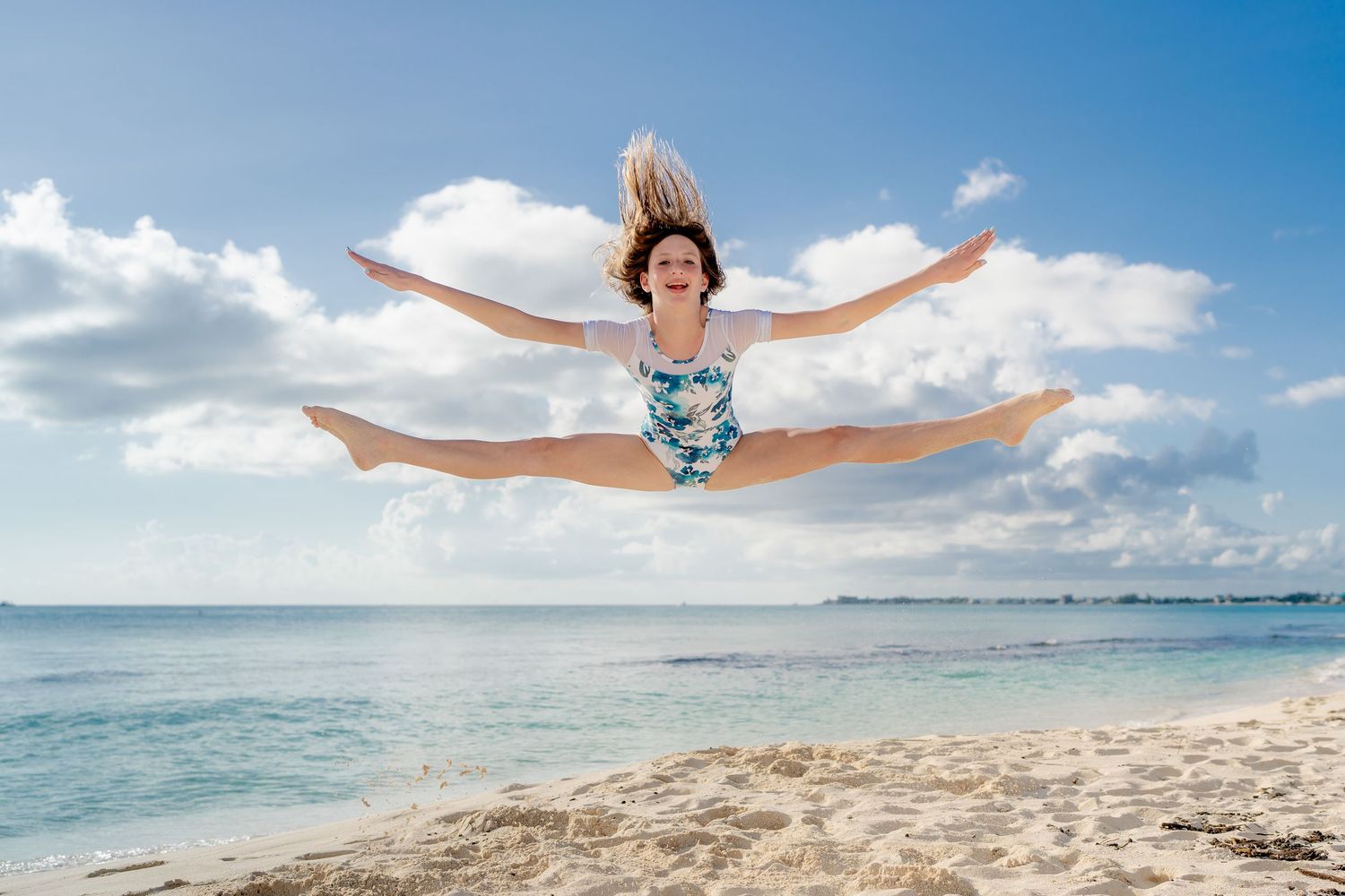 Person doing splits jump on tropical beach with blue sky and ocean.