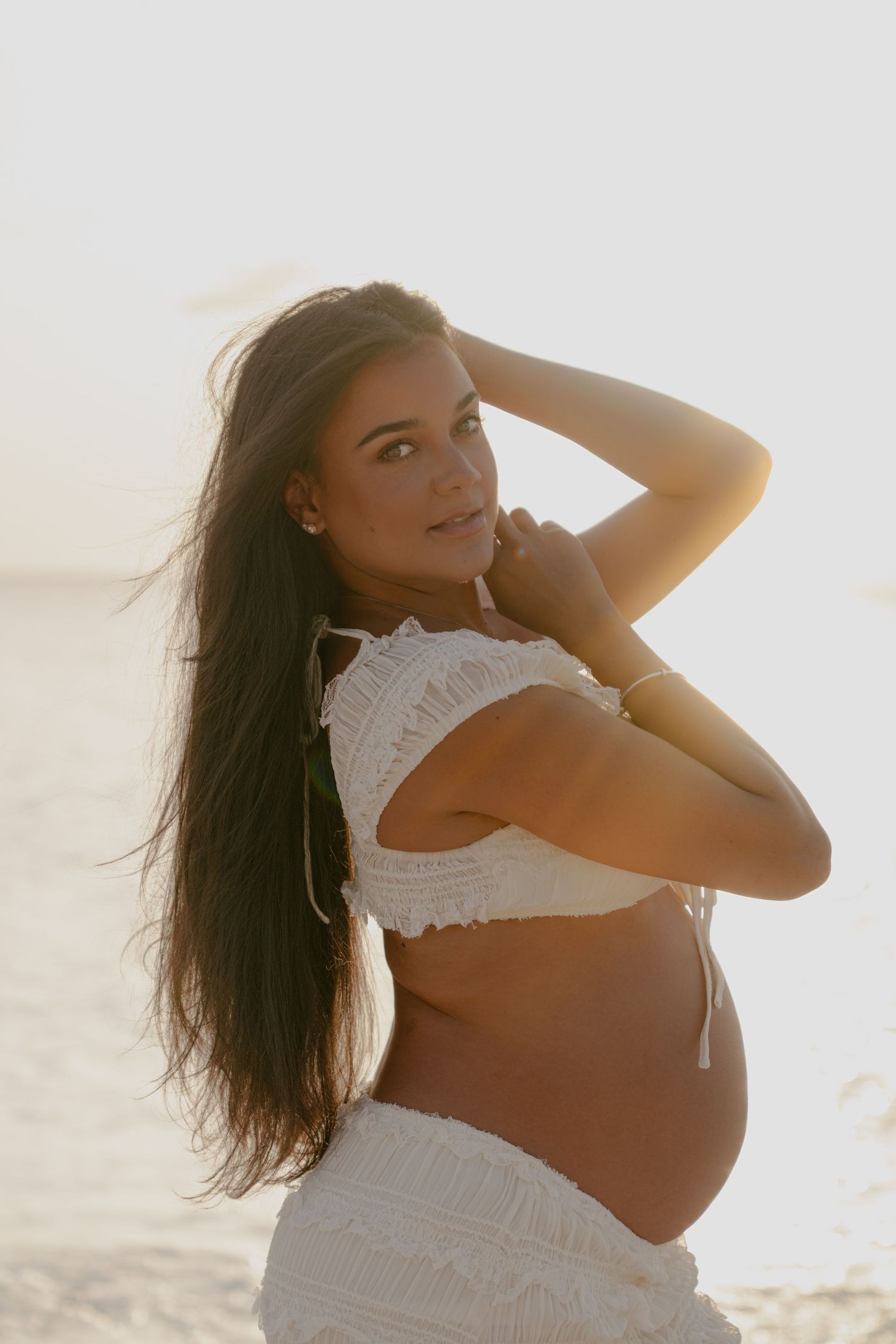 Expectant mother in white dress poses against bright beach backdrop.
