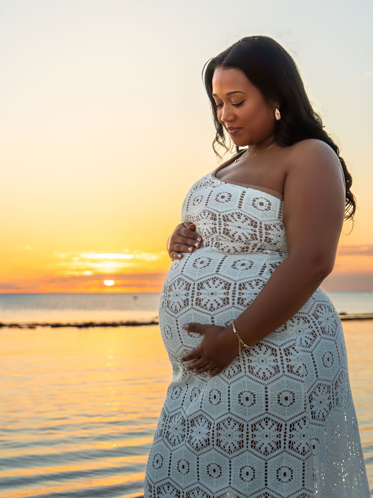 Pregnant woman in white lace dress cradling her baby bump at golden sunset by the ocean.