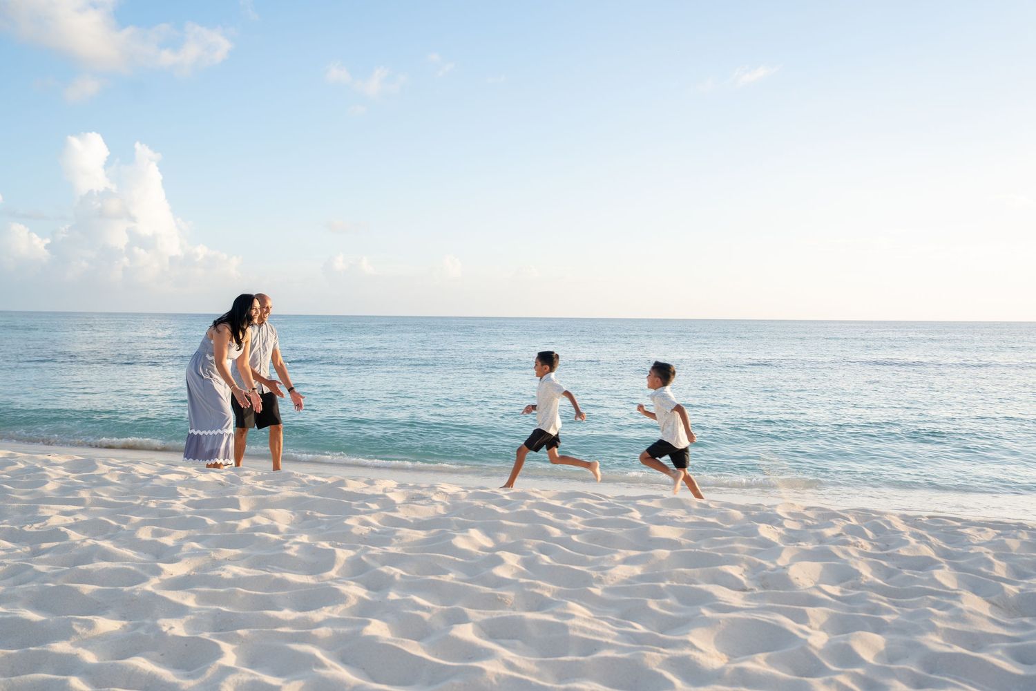 Family playing on a white sandy beach with calm turquoise ocean waters and clear blue sky in background.