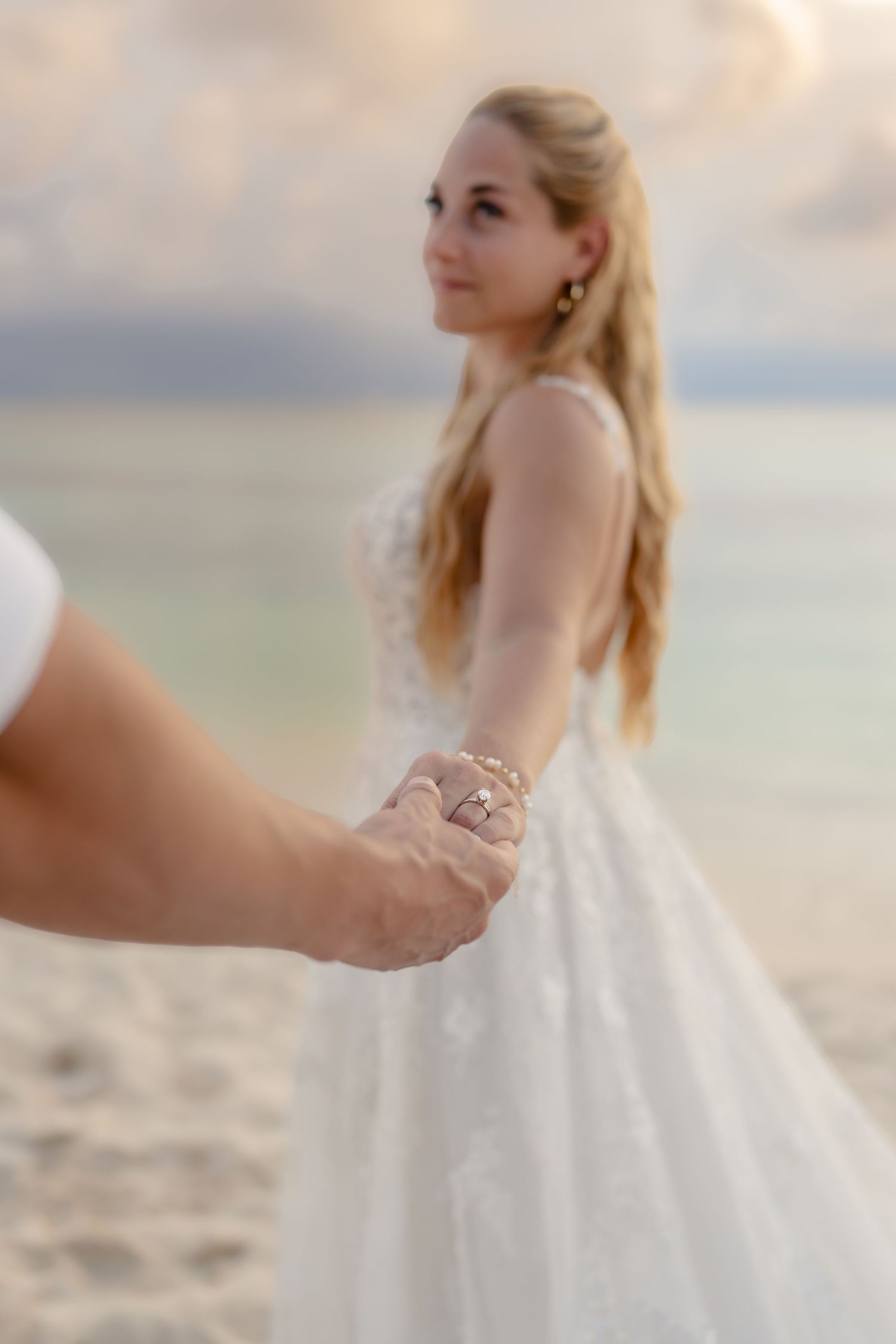 A bride in a flowing white lace gown poses gracefully on a sandy beach at sunset.