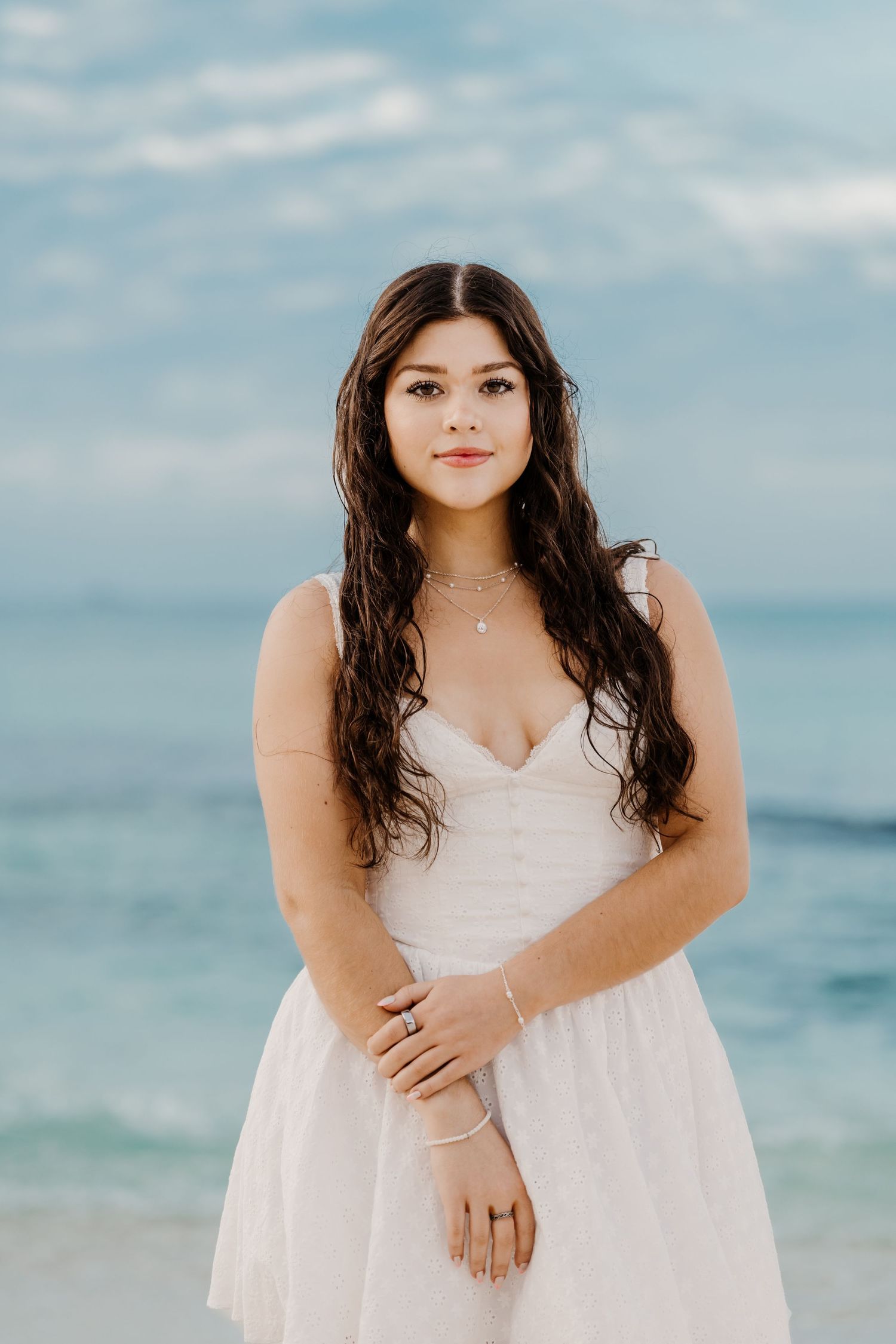 A person in a white dress poses on a beach with turquoise water and blue sky in the background.
