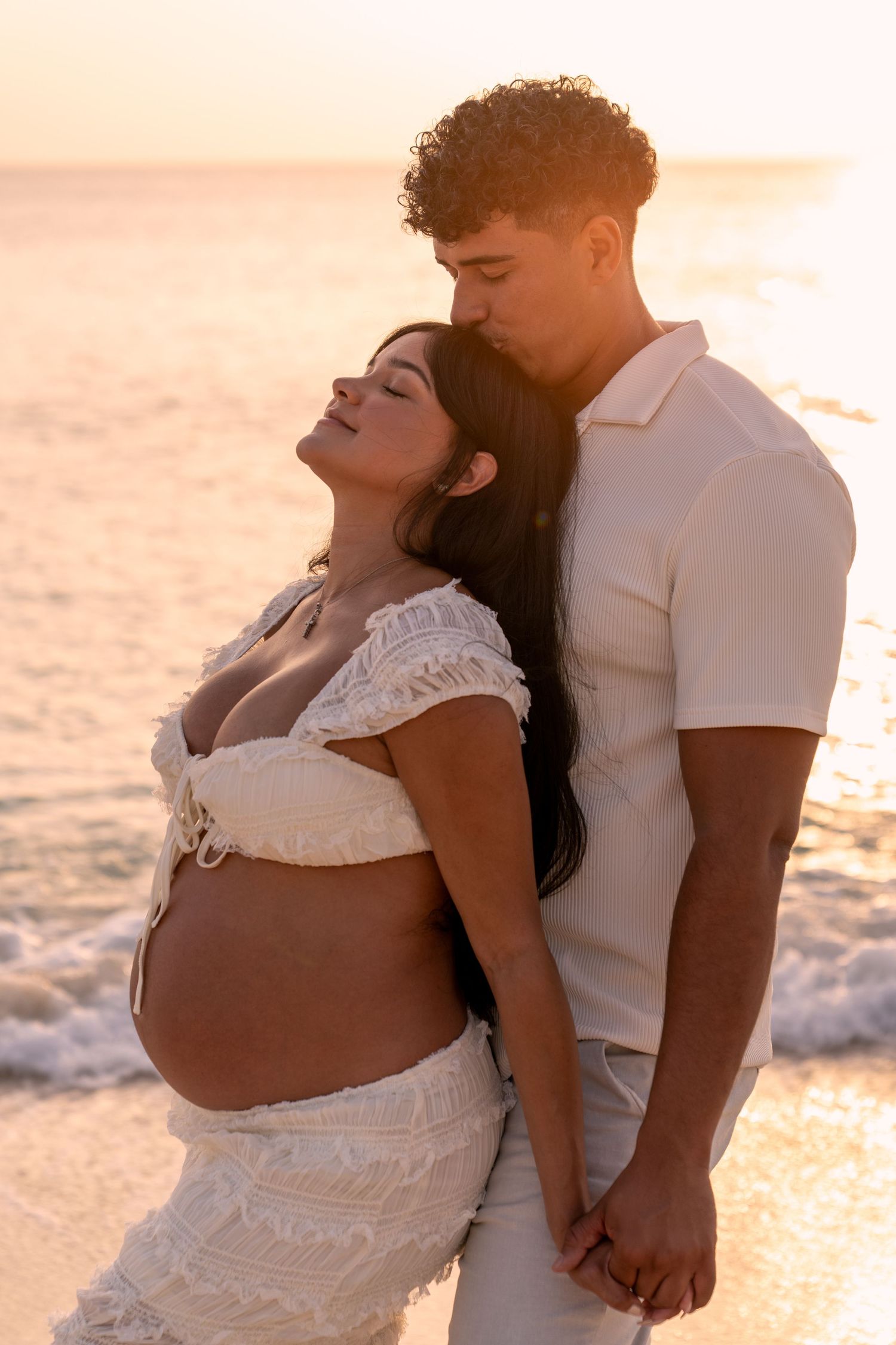 Romantic maternity photo of couple in white clothing standing on beach at golden hour.