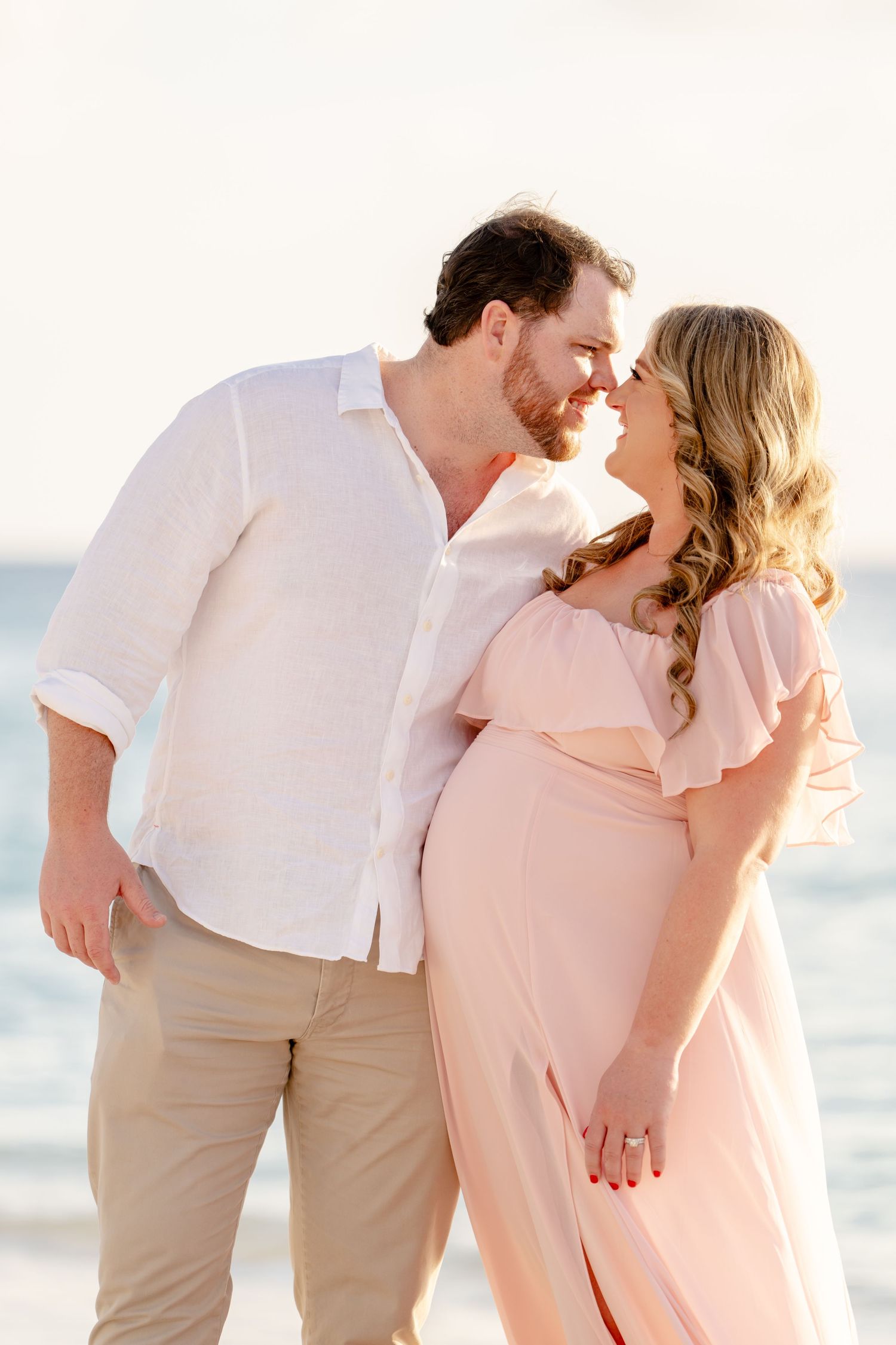 Couple in white and pink outfits sharing romantic moment on beach at sunset.