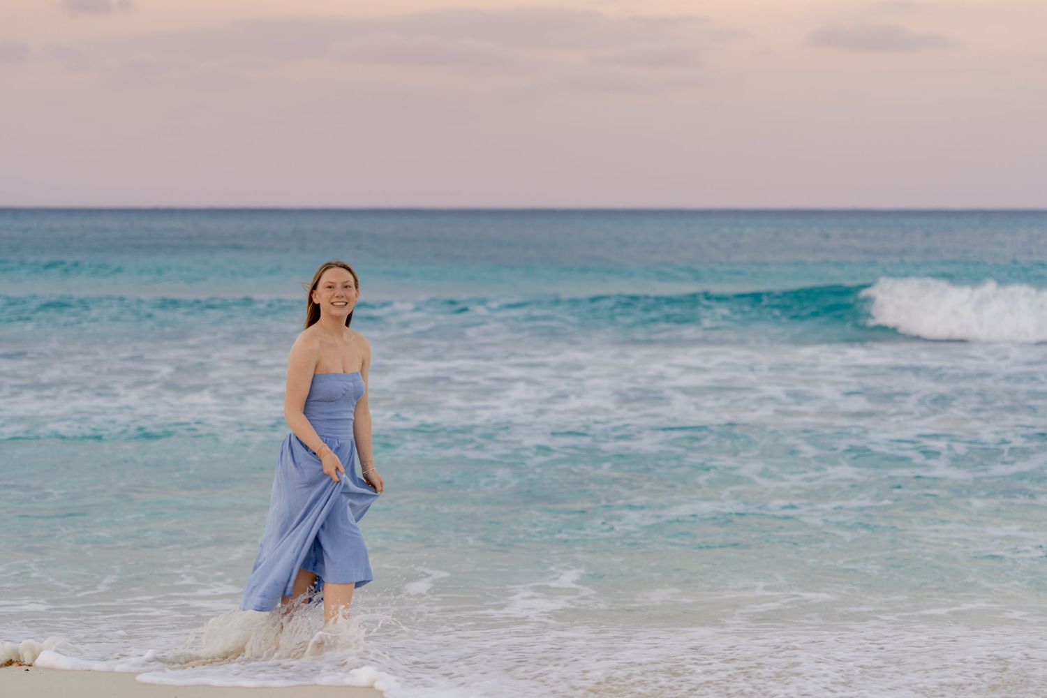 Person in blue dress running joyfully through shallow ocean water at golden hour.