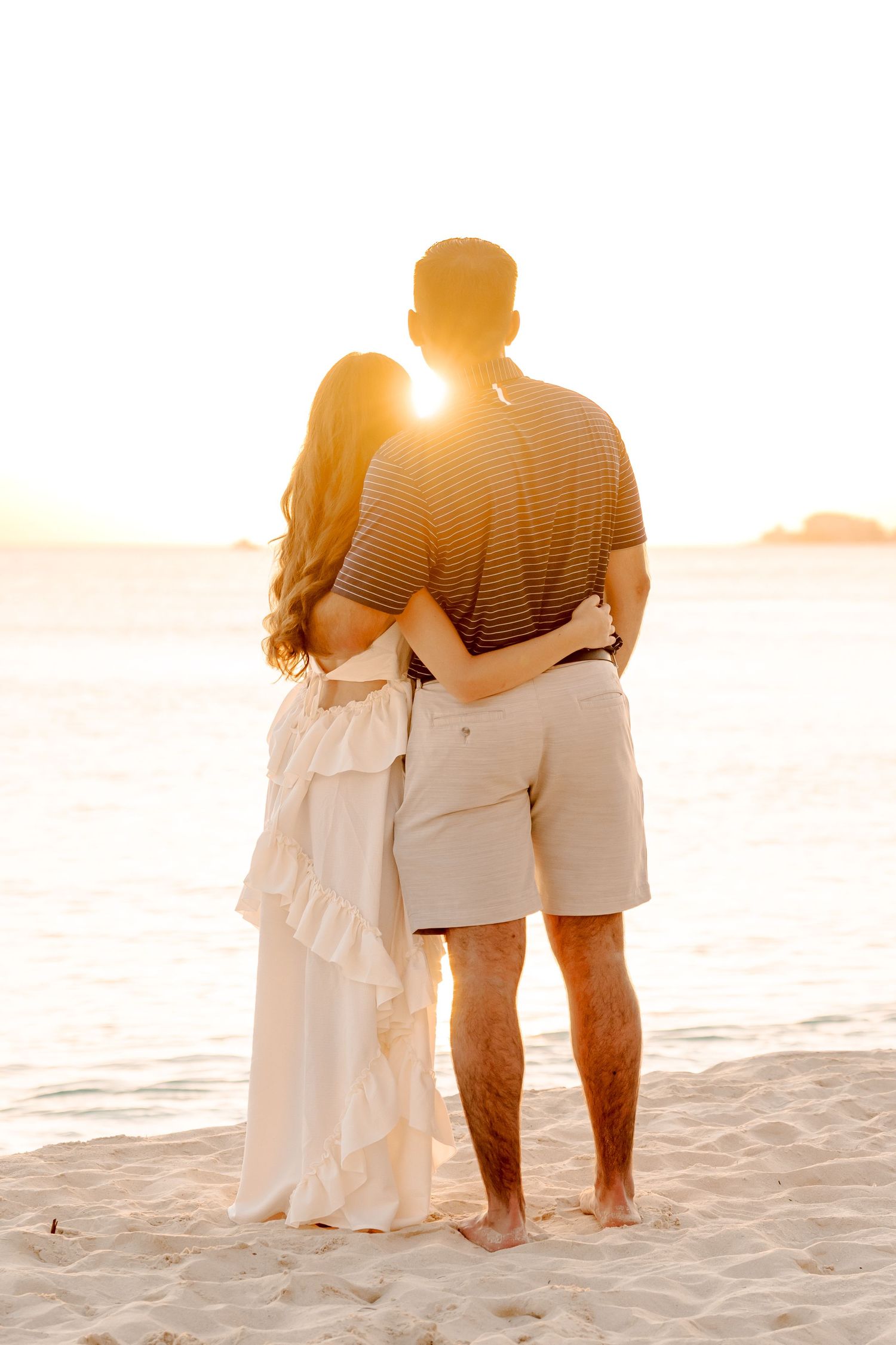 Couple standing together on sandy beach during sunset in white and casual attire.