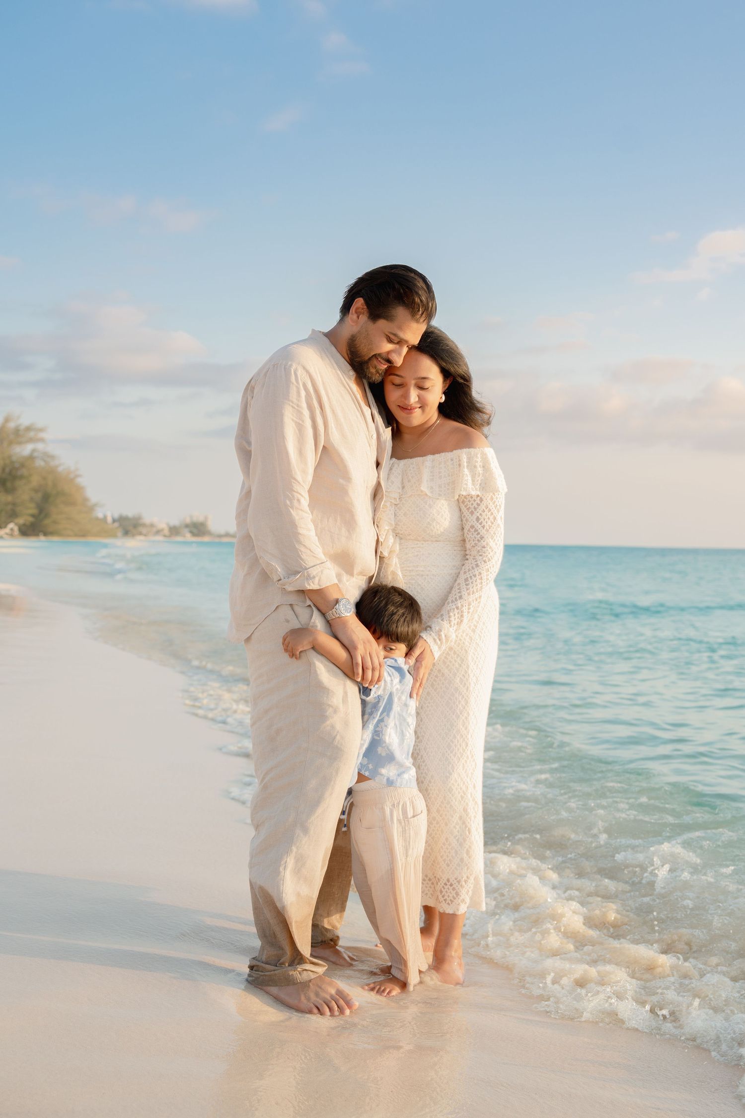 A happy family of three poses on a sandy beach, with parents embracing their young child near the ocean waves.