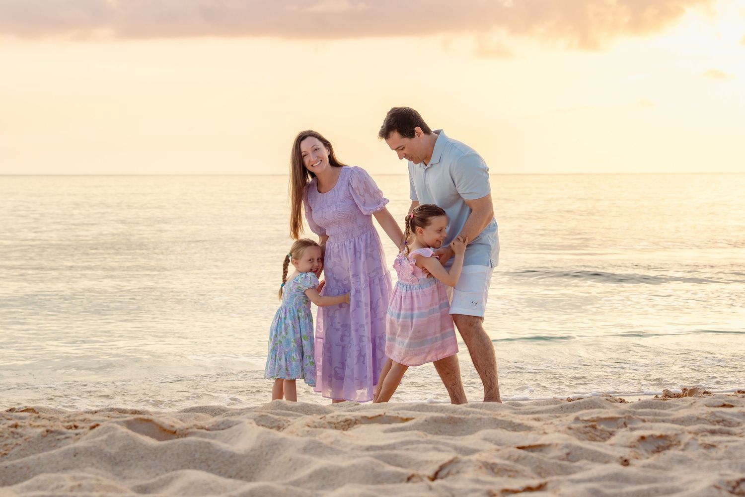 Family of four enjoying a golden sunset beach walk, with two young girls in matching floral dresses on sandy shore.