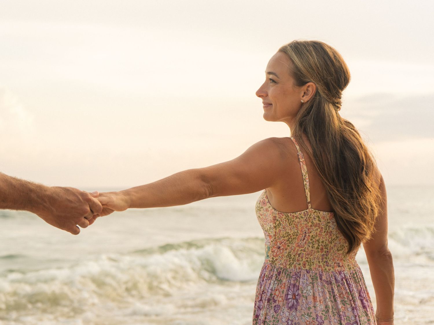 Woman in floral sundress holding hands with partner on a serene beach at sunset with soft waves behind her.