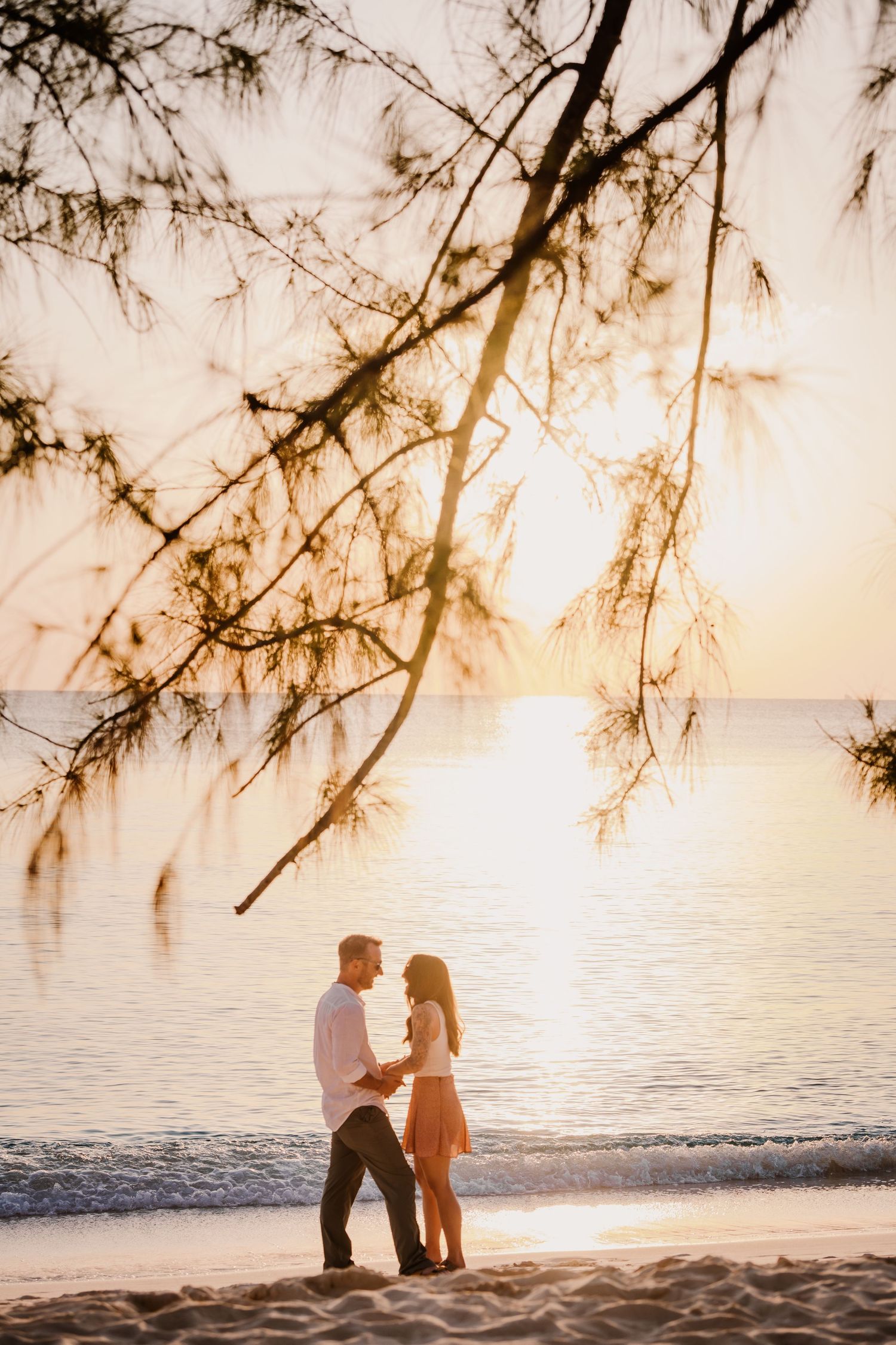 A couple stands together on a beach at sunset with overhanging tree branches framing the romantic scene.