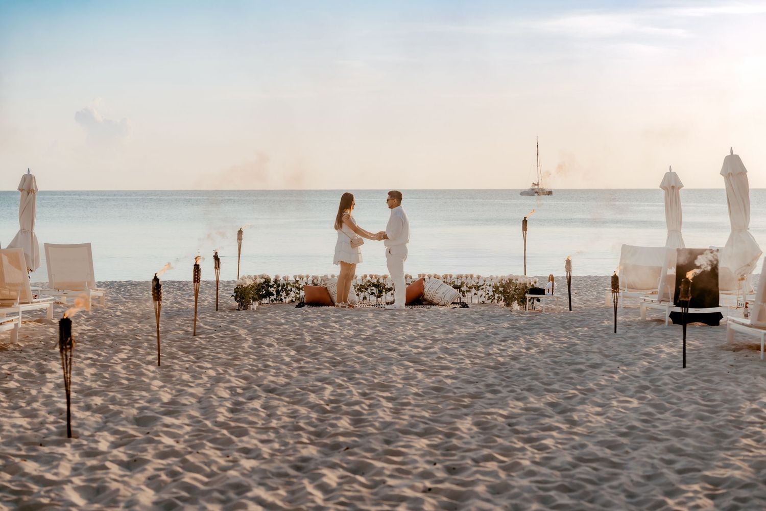 A panoramic view of a pristine sandy beach with tiki torches and beach furniture at sunset.