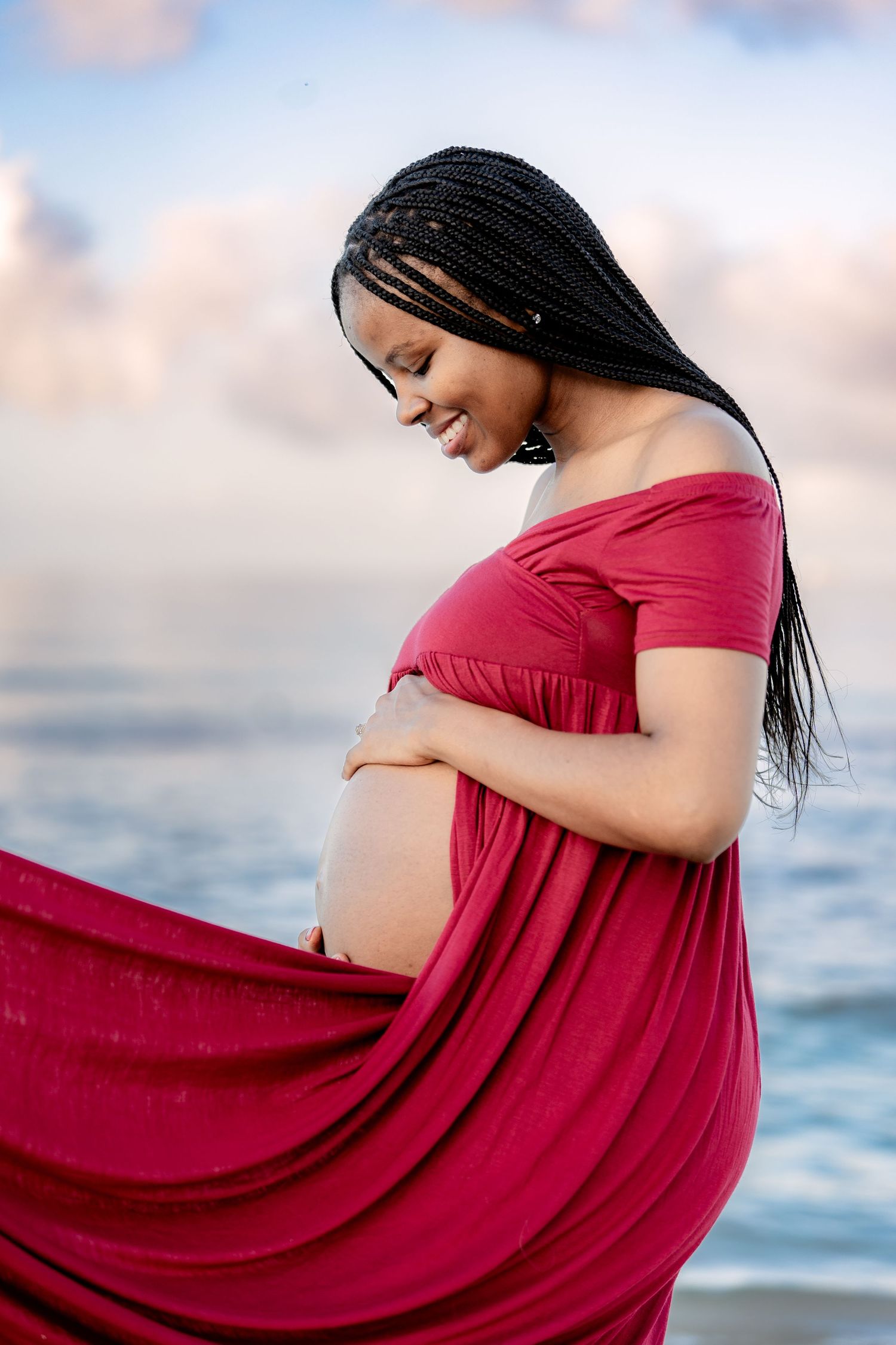 Expectant mother in flowing red dress poses gracefully by ocean during sunset maternity shoot.