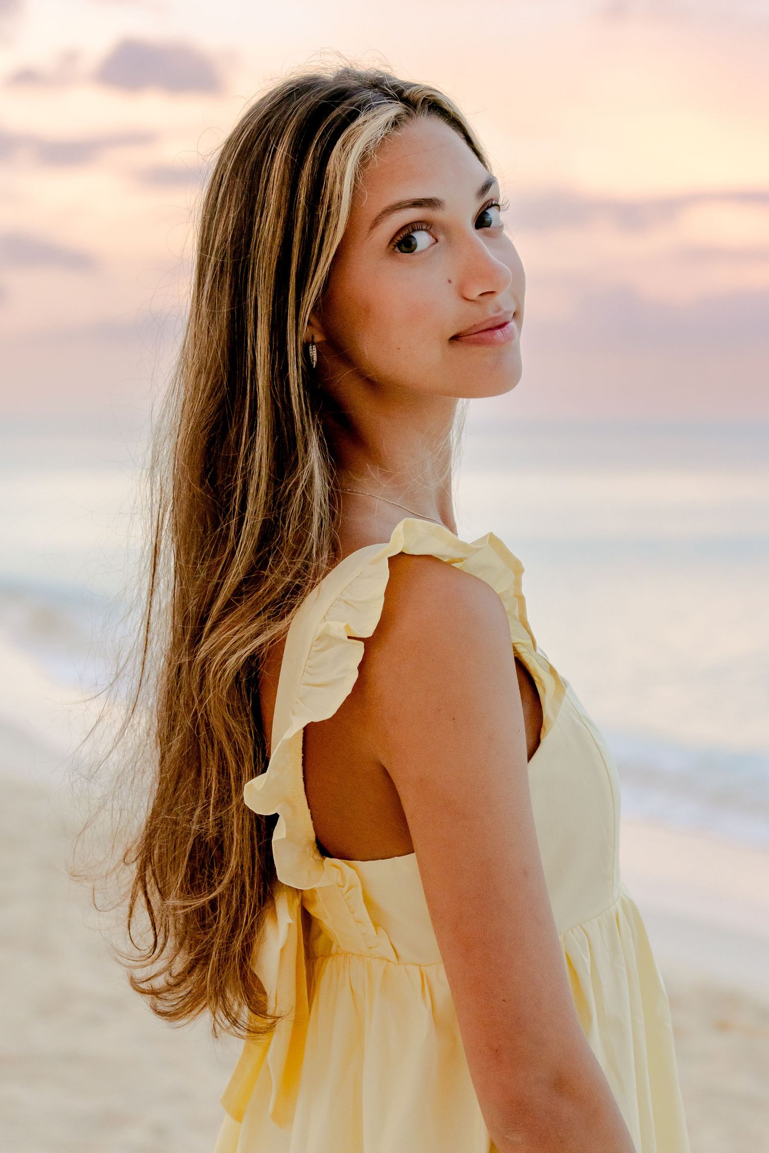 Someone wearing a pale yellow ruffled dress poses against a pastel sunset sky at the beach.
