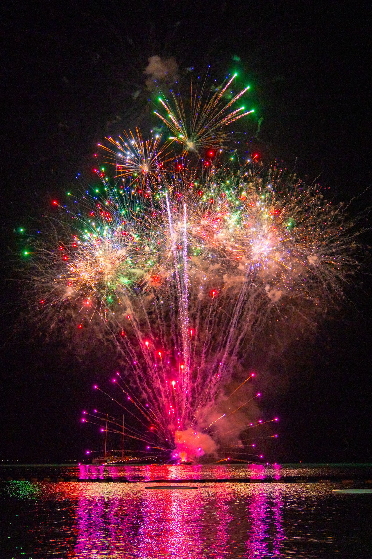 Vibrant fireworks display reflecting on water with red, green and pink colors illuminating the night sky.