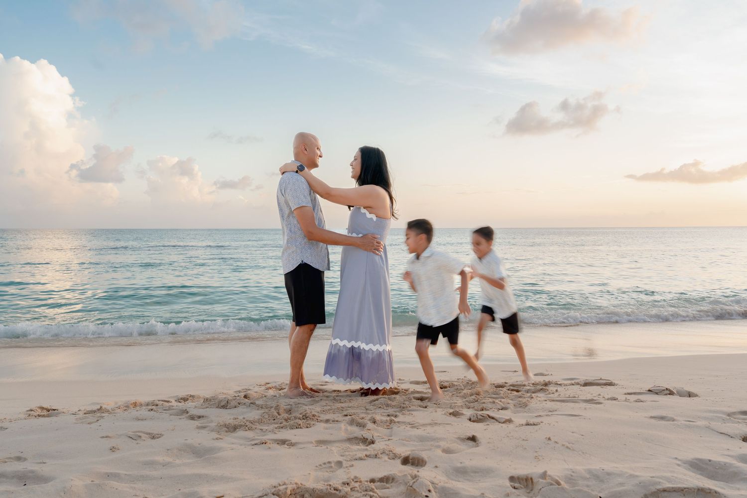 Family of four enjoying a serene beach sunset, parents embracing while two boys play on sandy shore.