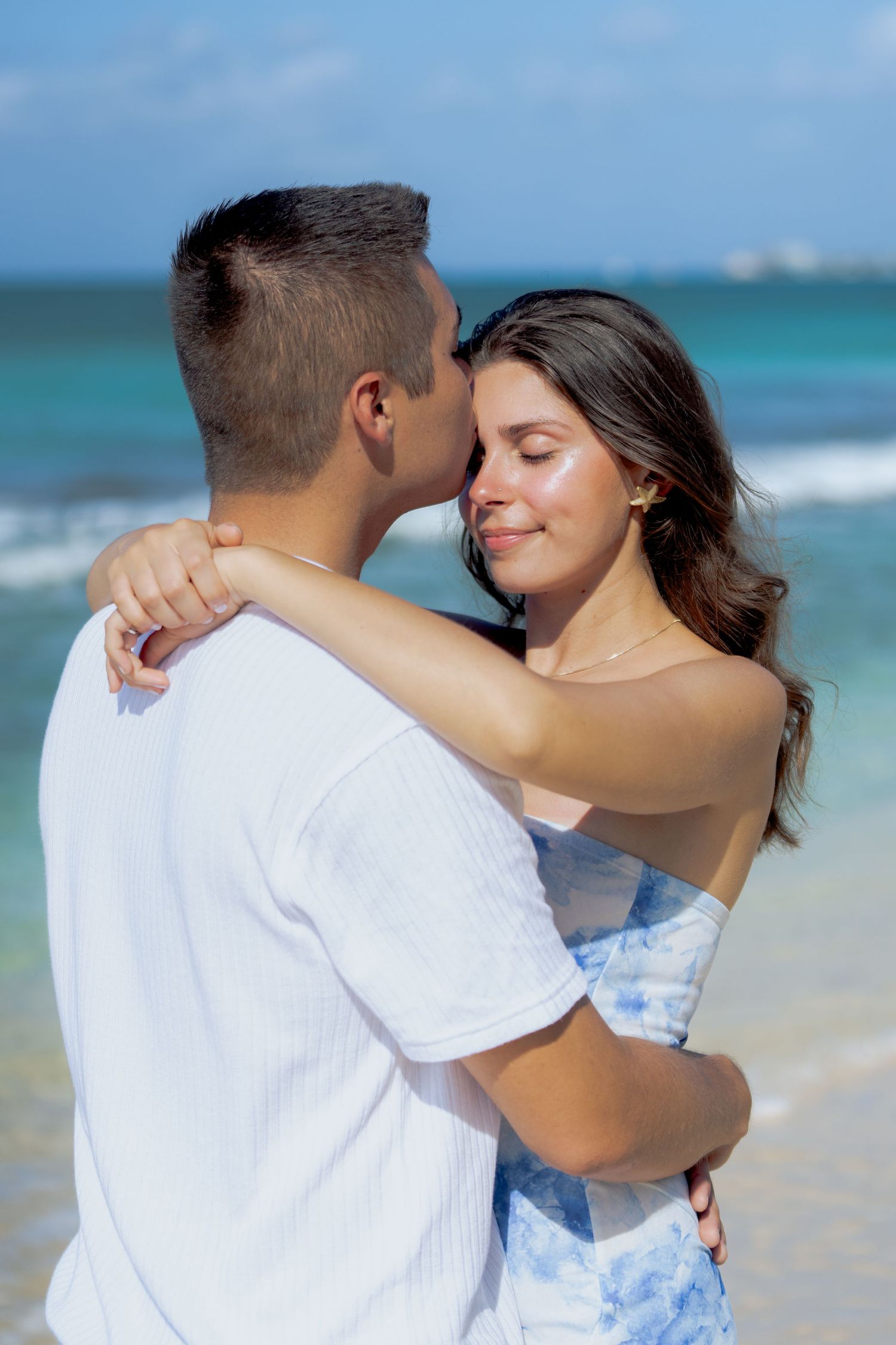 Romantic couple embracing on a tropical beach with turquoise waters and blue sky in the background.