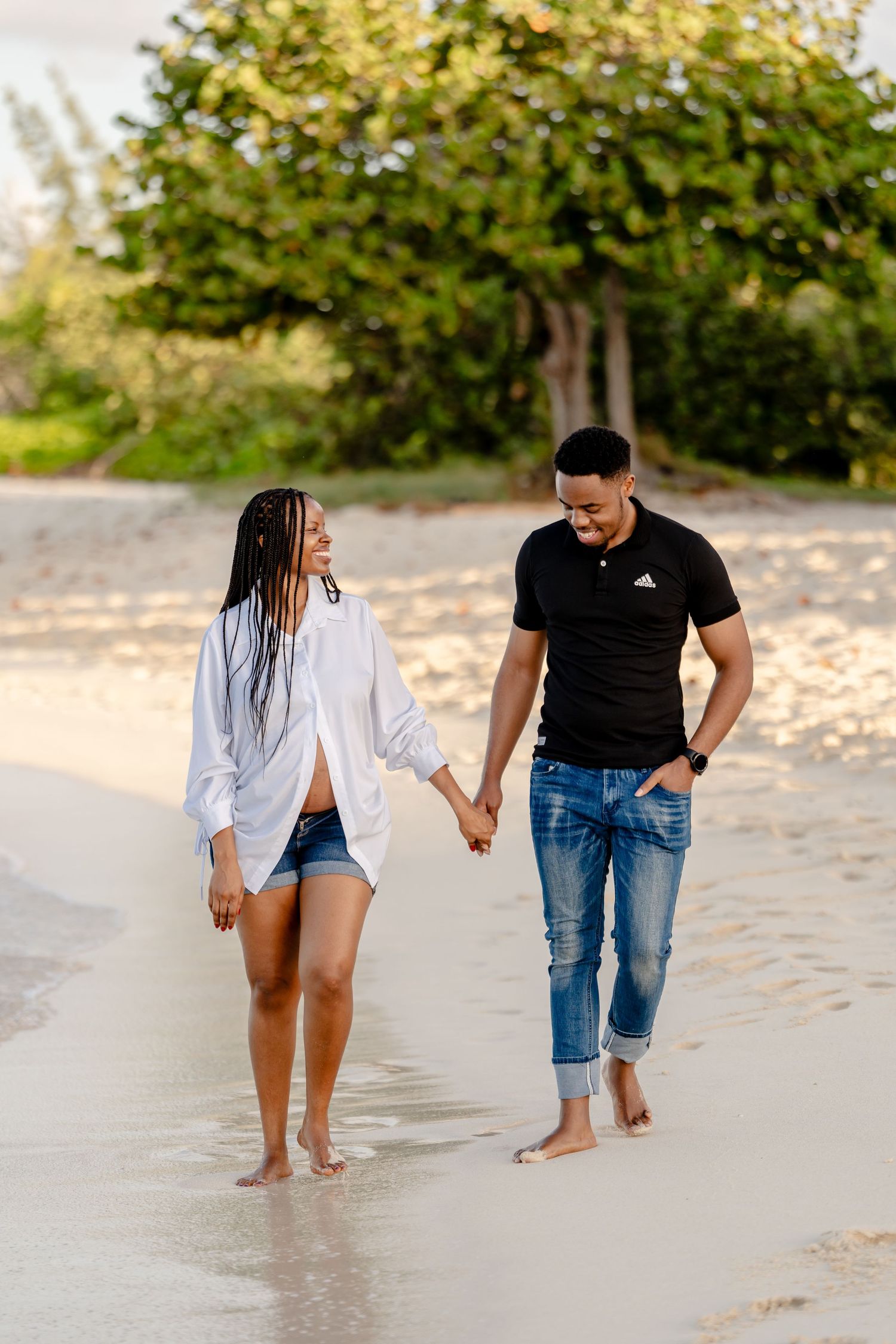 Couple walks hand in hand along sandy beach wearing casual outfits during sunset.