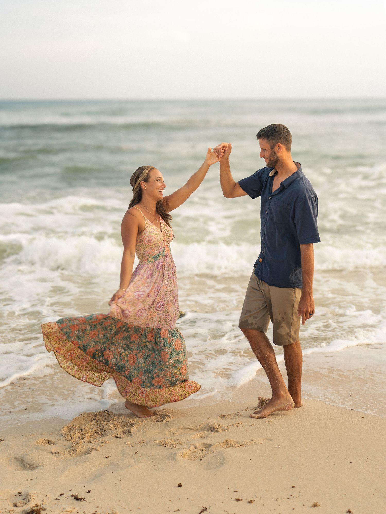 Couple dancing barefoot on a sandy beach at sunset, woman in floral dress twirling.