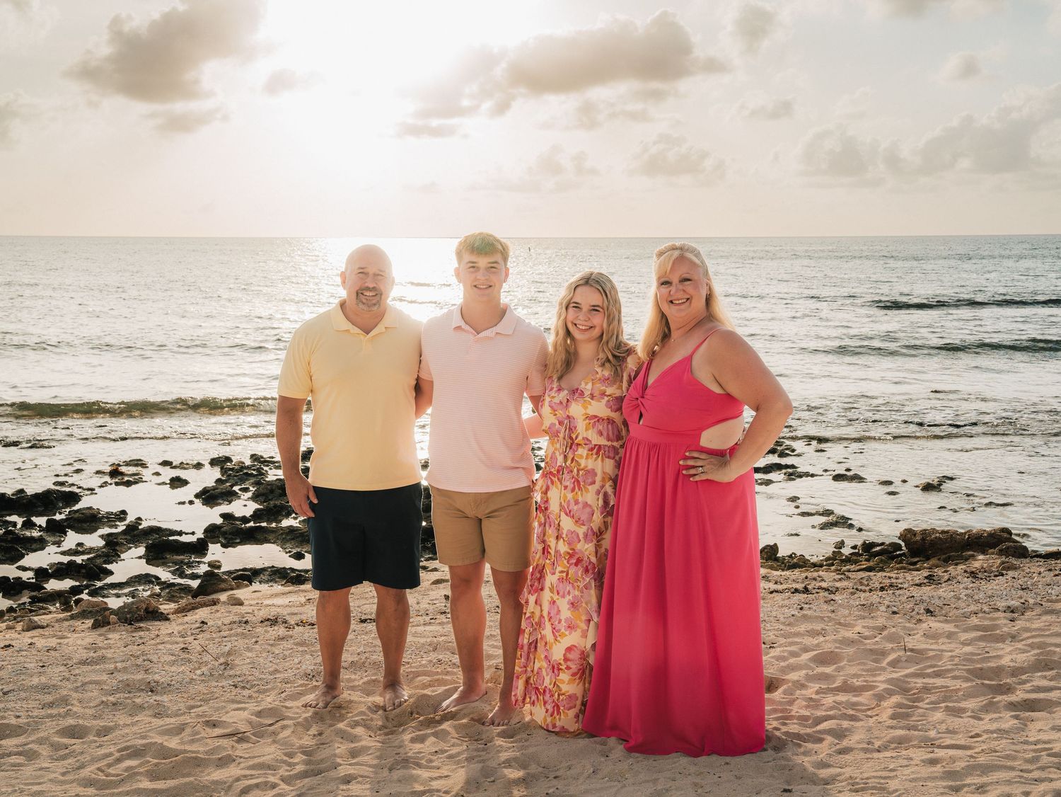 Family of four smiling together on a sandy beach at sunset with ocean waves in the background.