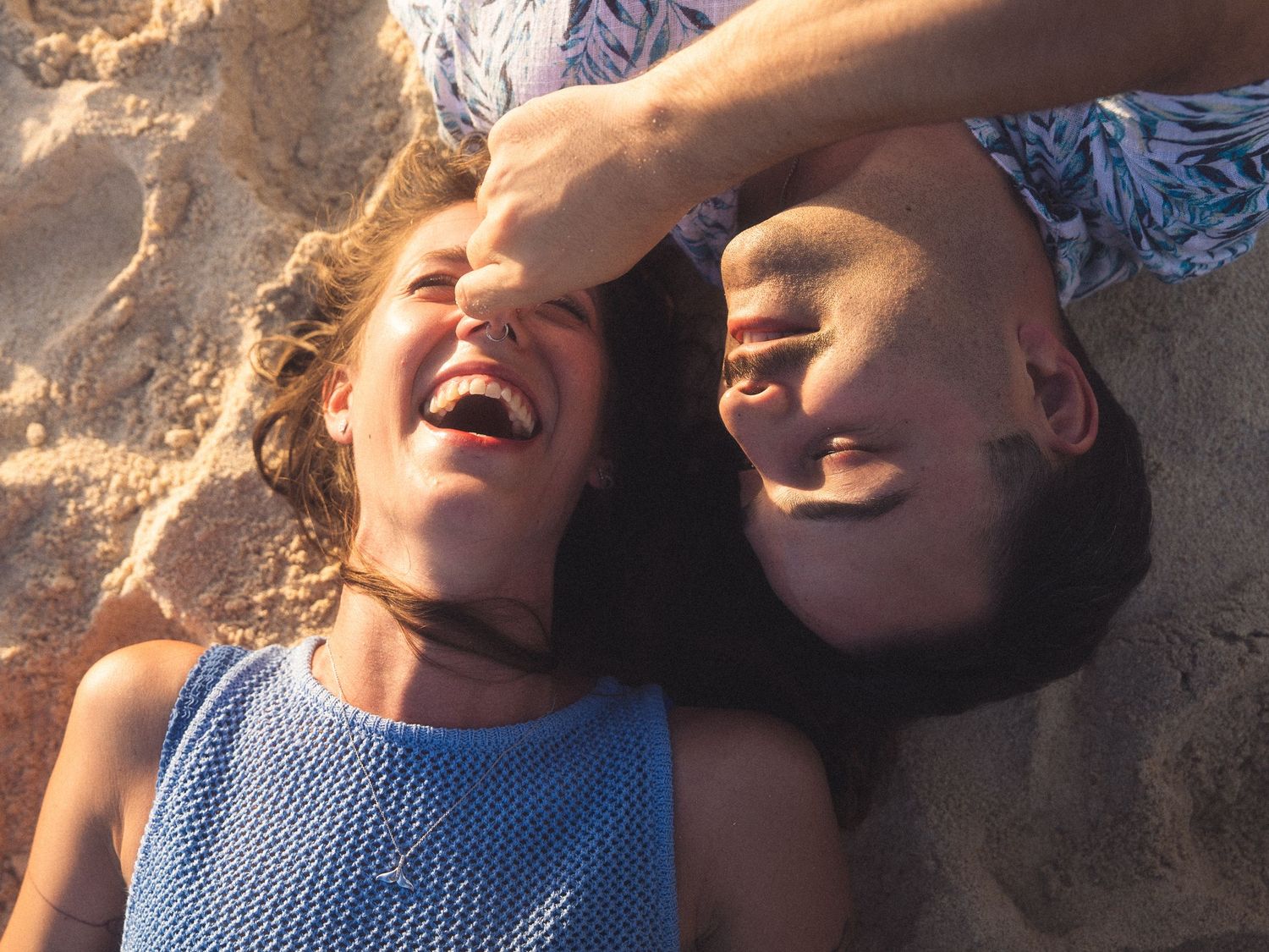 Two women laughing joyfully while lying on sandy beach together.