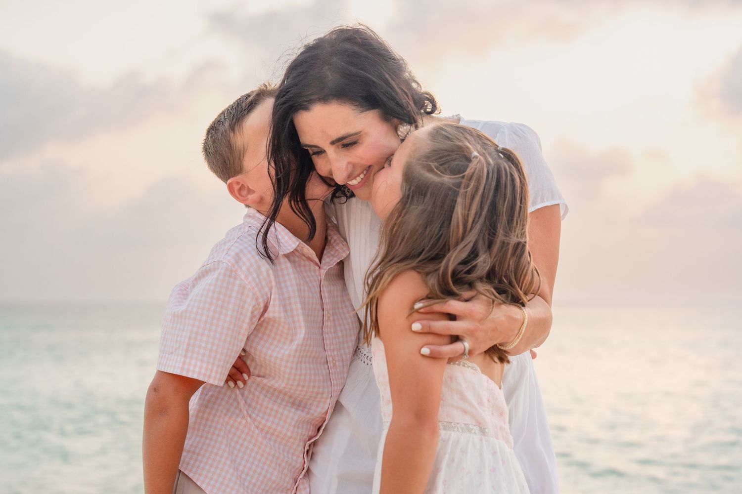 A mother shares a joyful beach embrace with her two young children at sunset by the ocean.