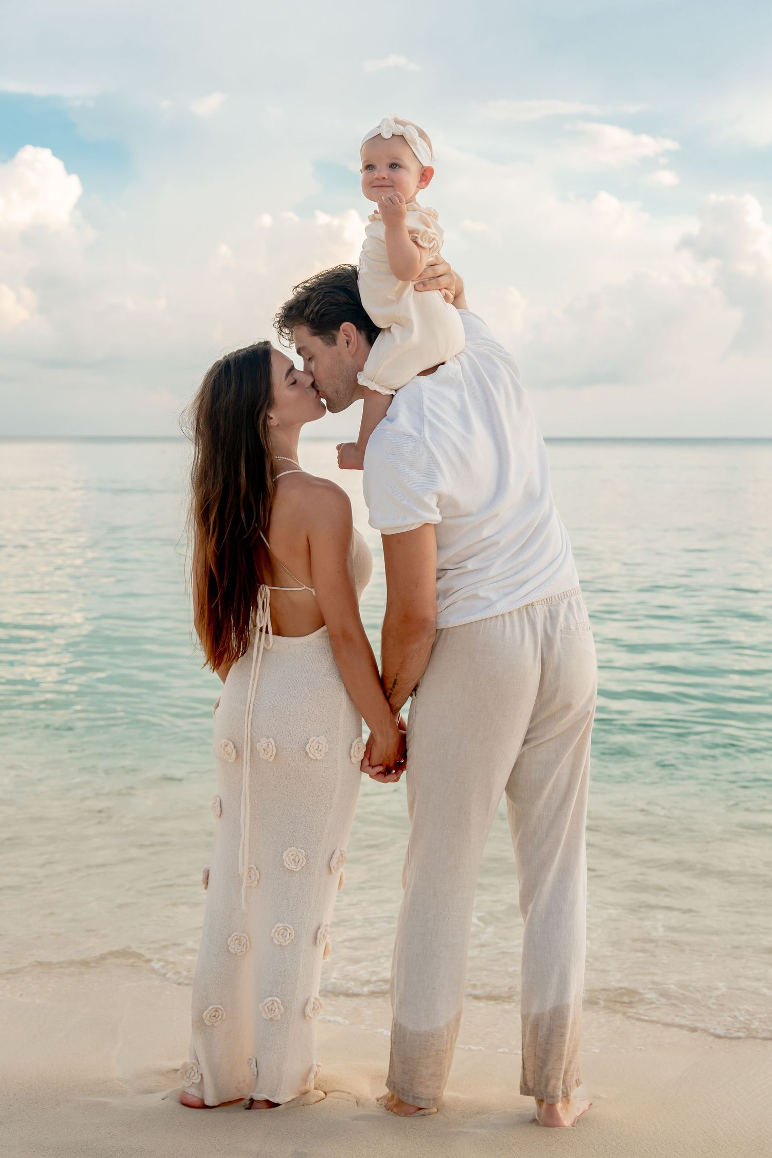 A couple kisses on a tropical beach while holding their baby girl, all dressed in white outfits.