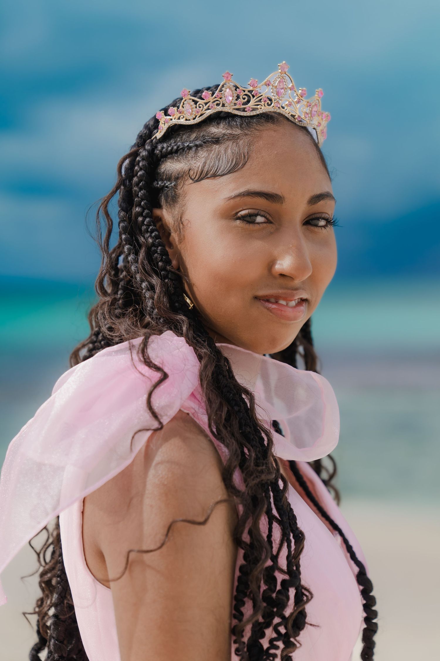Person wearing pink dress and tiara poses on beach with turquoise waters in background.