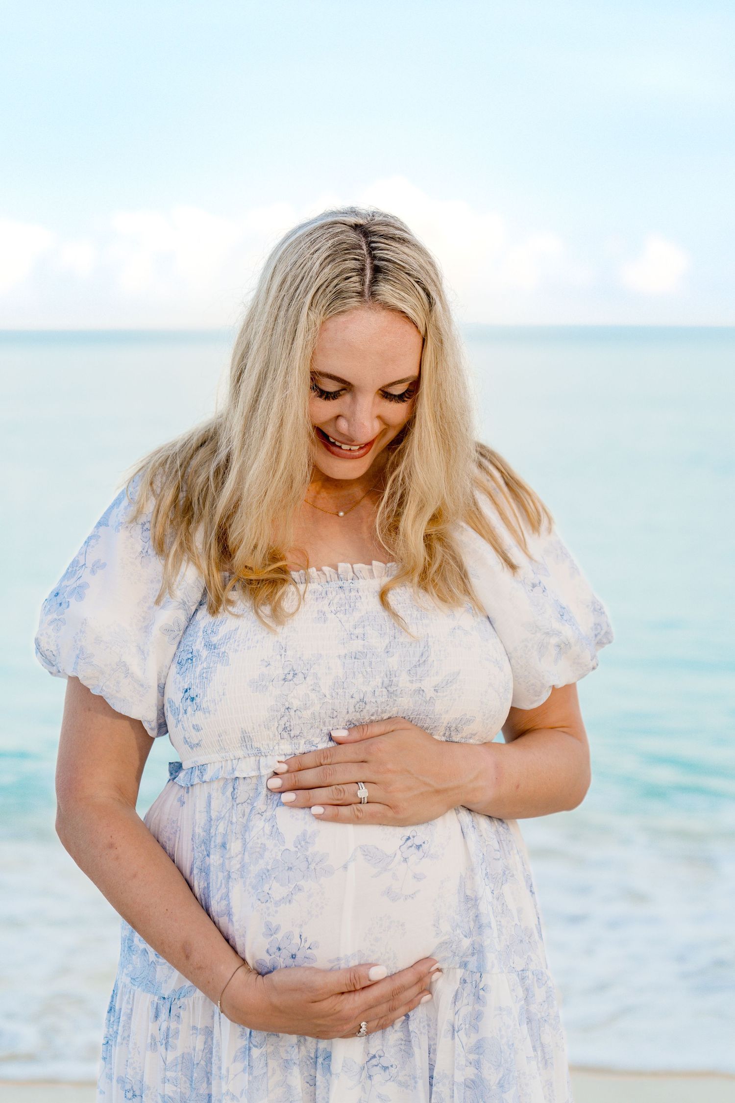 Woman in floral dress cradling baby bump on tropical beach.