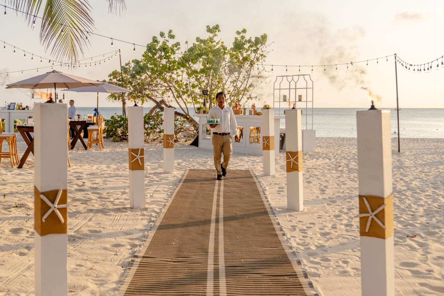 Beach wedding setup with lanterns and decorations along a sandy pathway at sunset.