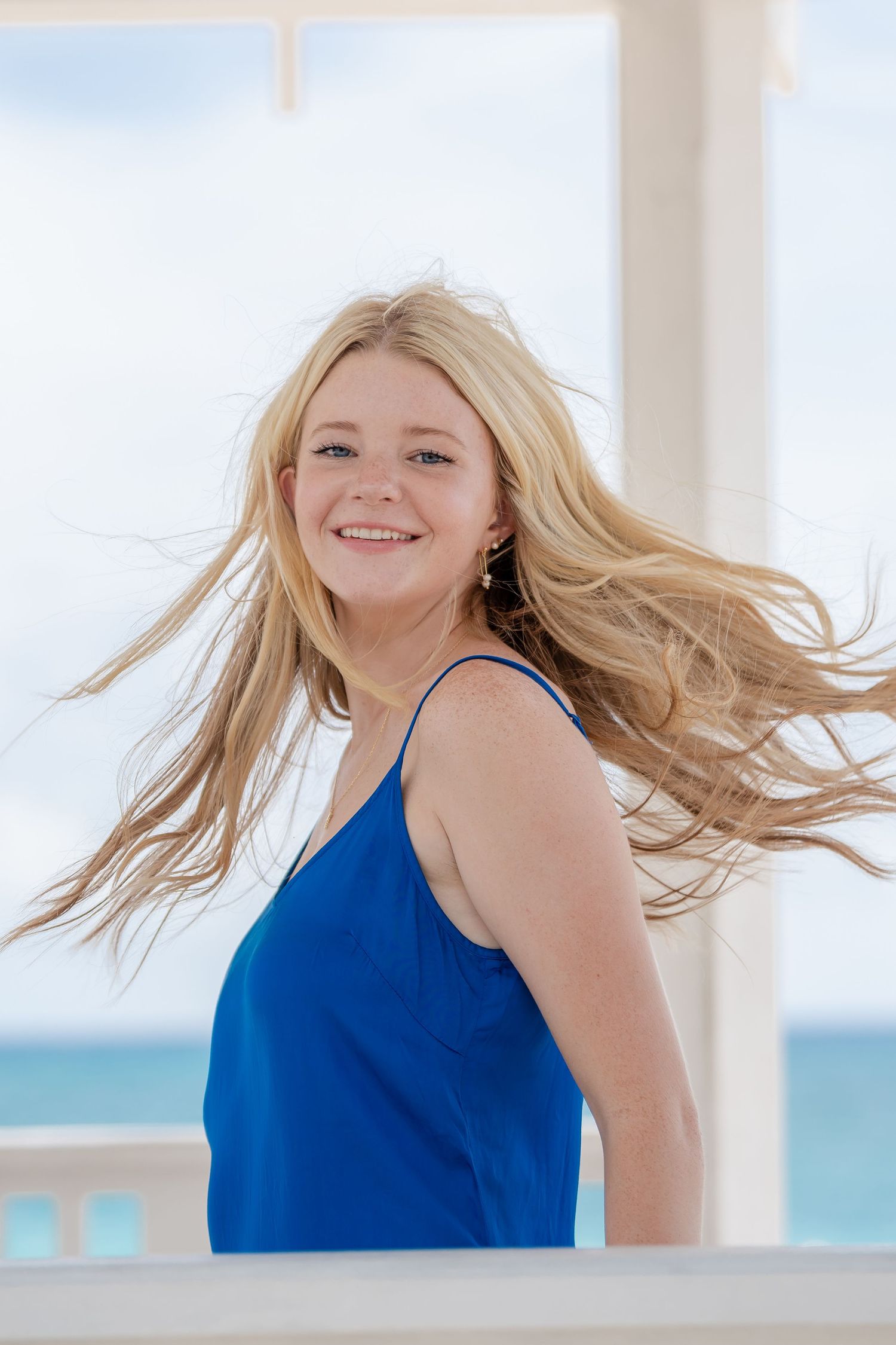 Woman in blue sleeveless top poses against bright coastal background with windswept hair.