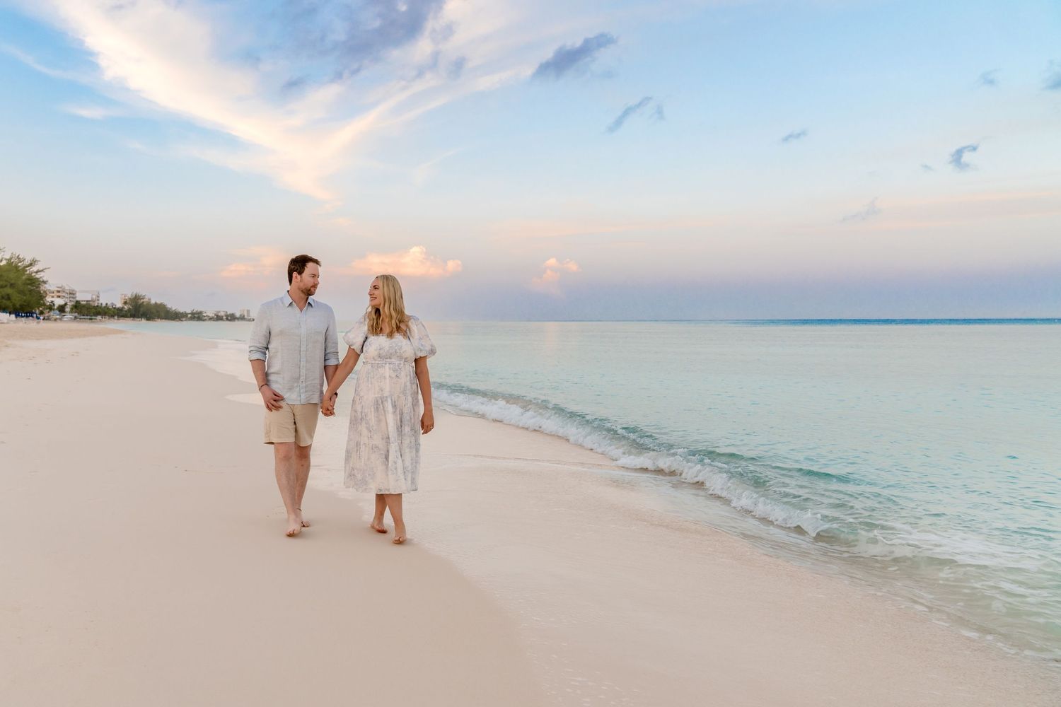 A couple walks hand in hand along a pristine white sand beach at sunset with turquoise waters in the background.