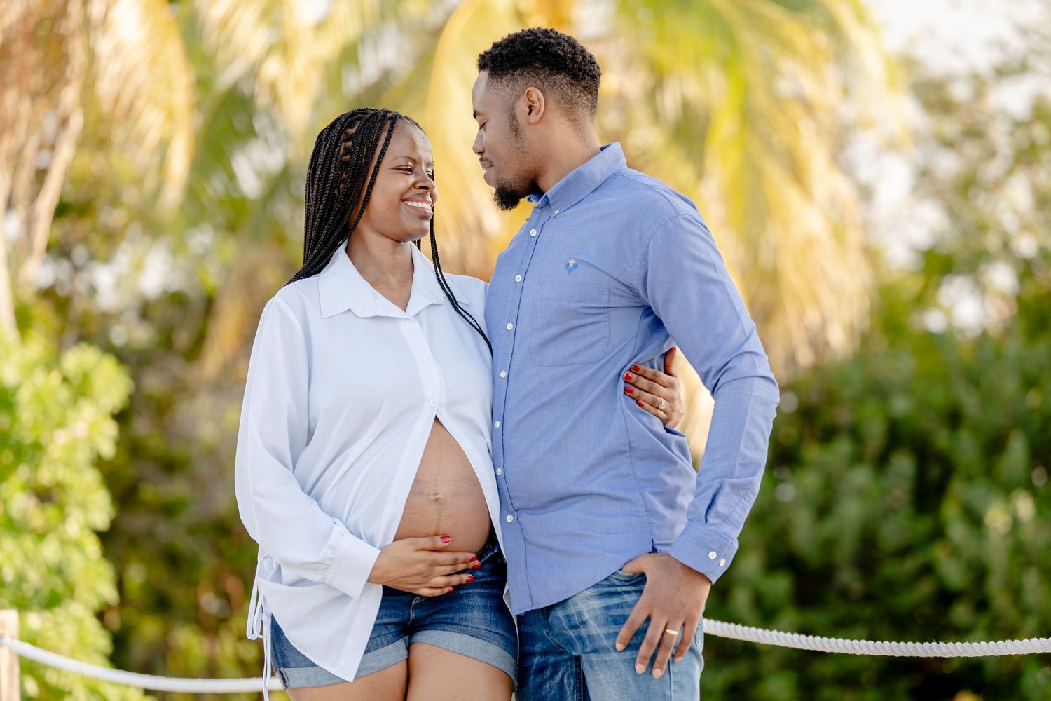 Couple embraces during outdoor maternity photoshoot with palm trees in background.