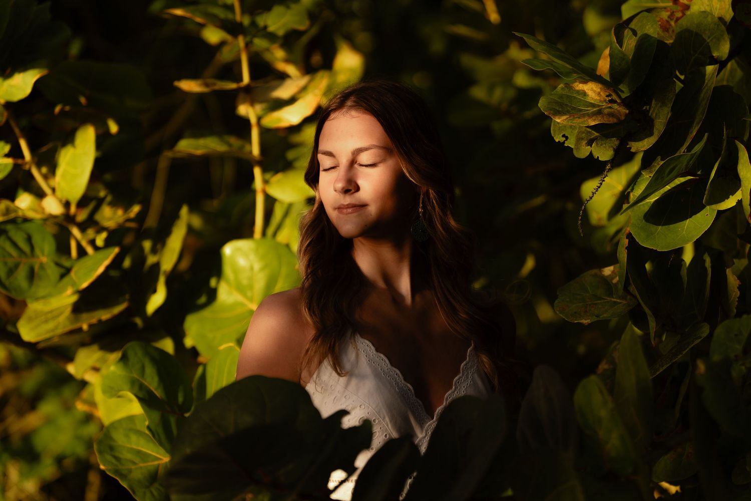 Woman in white dress poses thoughtfully against lush green foliage in golden sunlight.