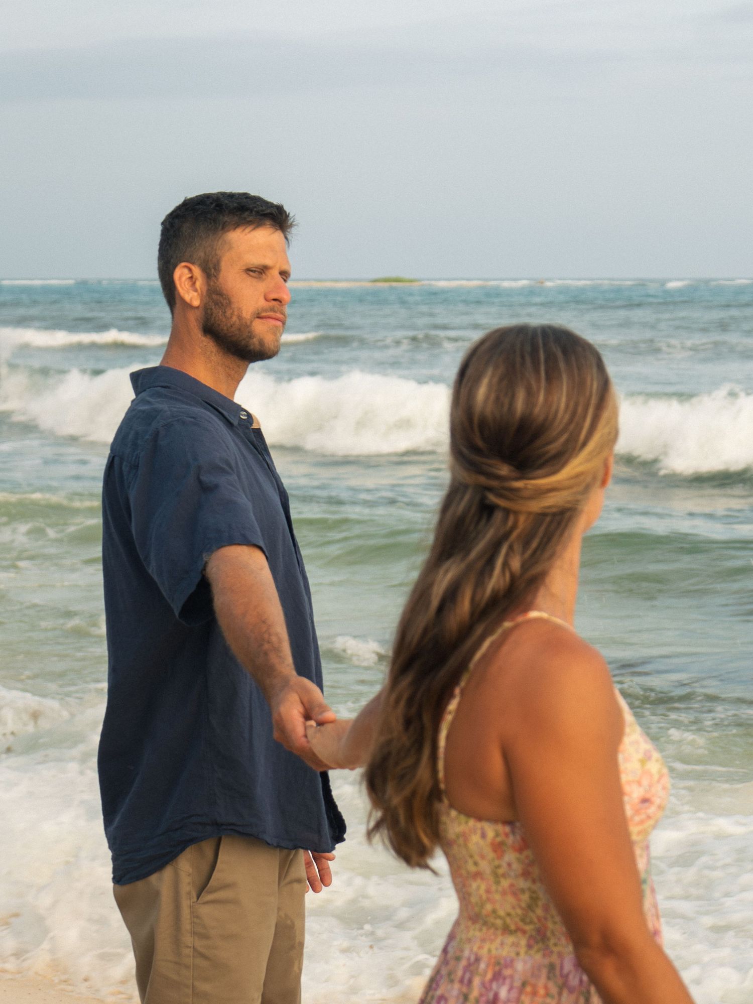 Couple holding hands on a tropical beach with ocean waves, man in navy shirt and woman in floral dress facing each other.