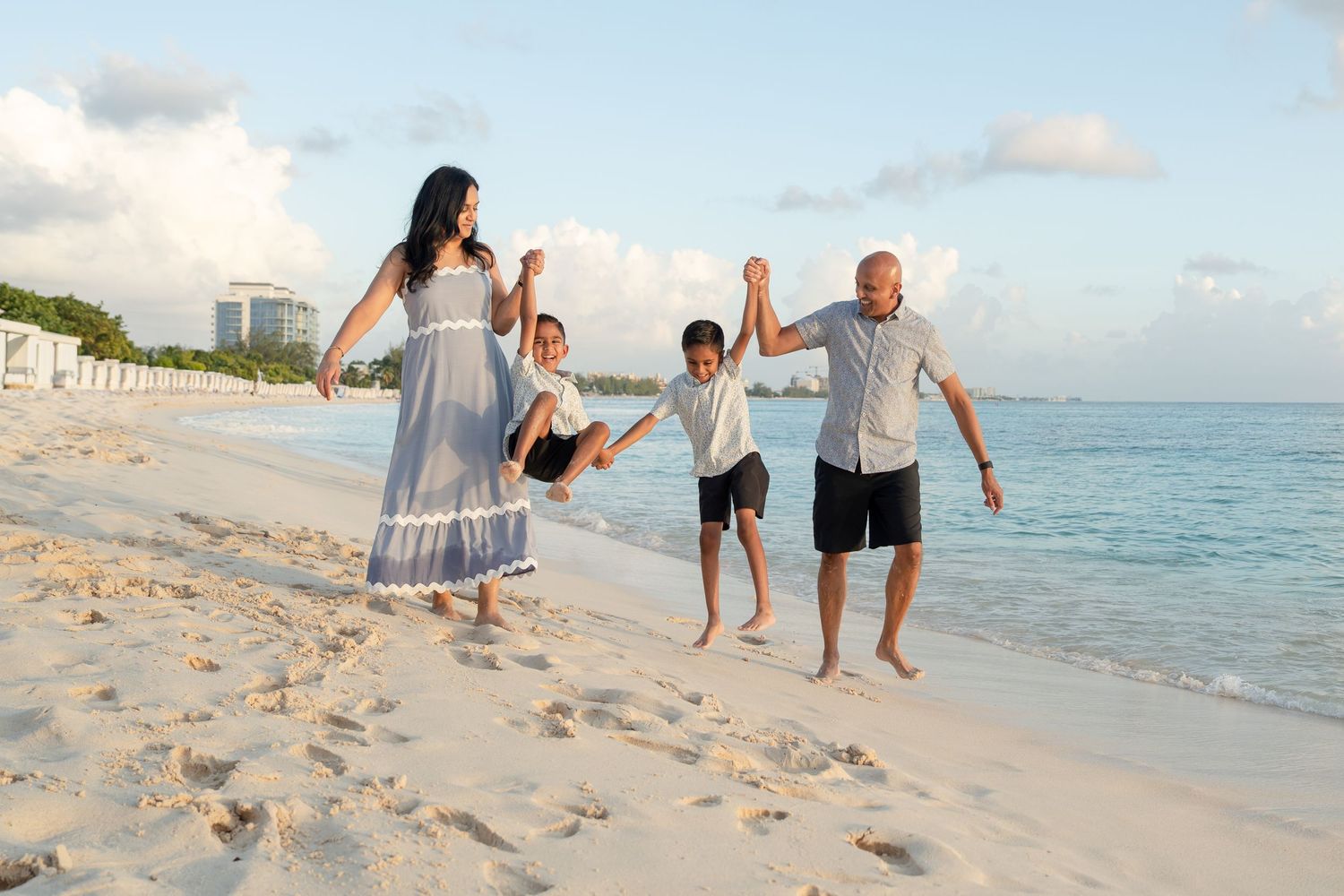 Family of four joyfully swinging children on a sunny white sand beach with turquoise water in the background.