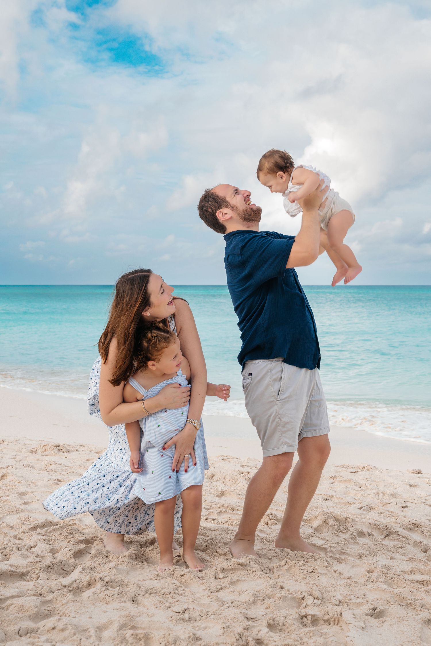 A family enjoys a playful moment together on a tropical beach with turquoise waters and cloudy blue skies.