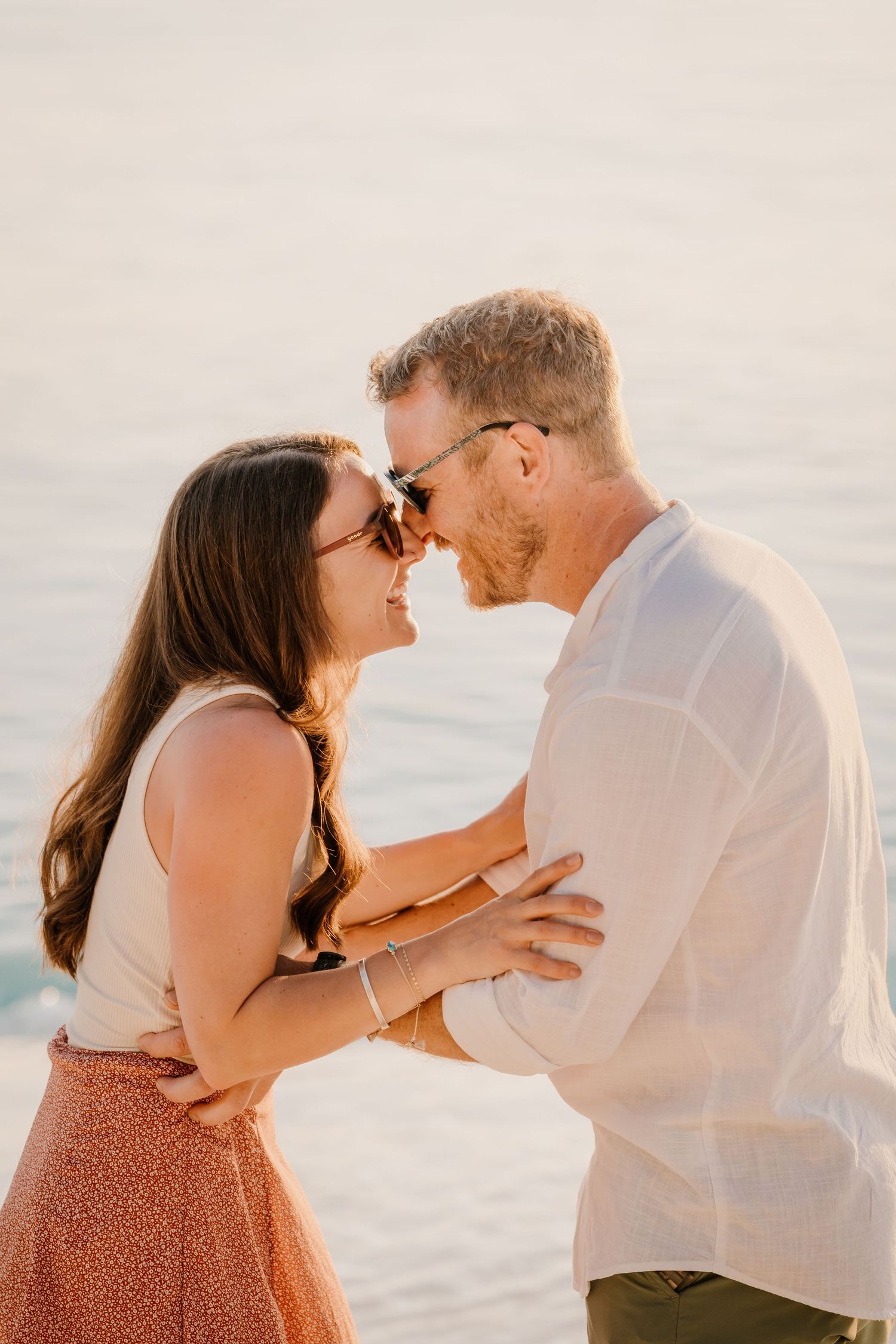 A romantic couple sharing an intimate moment at sunset by the ocean, wearing white and coral colored outfits.