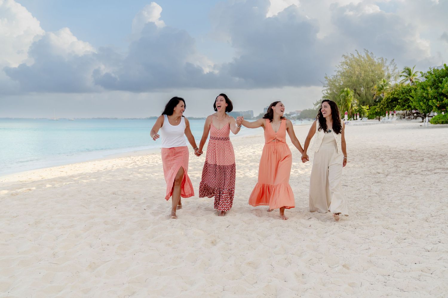 Friends in coordinated coral and white dresses walk together on a scenic beach.