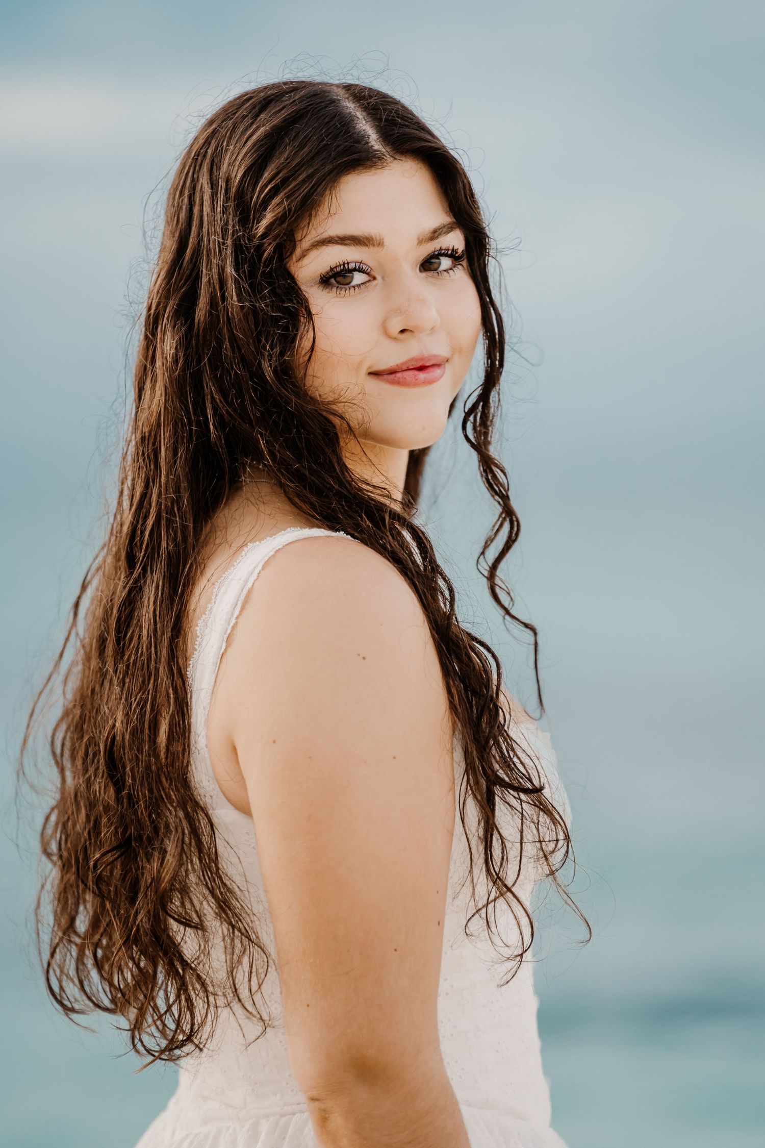A person in a white dress poses against a blue ocean backdrop during a beach photoshoot.