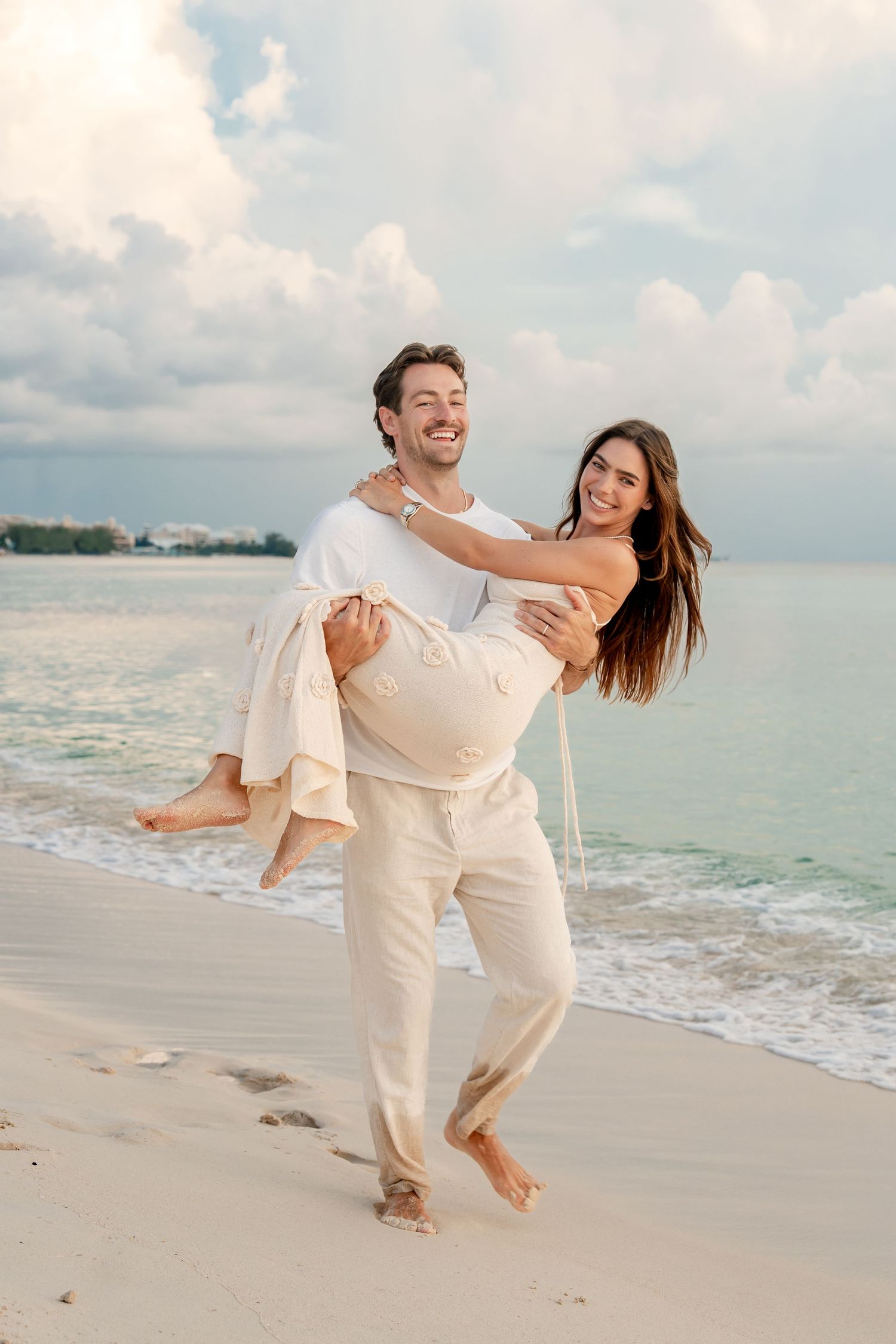 Happy couple on a beach, man carrying woman in his arms, smiling, with ocean waves and cloudy sky behind them.