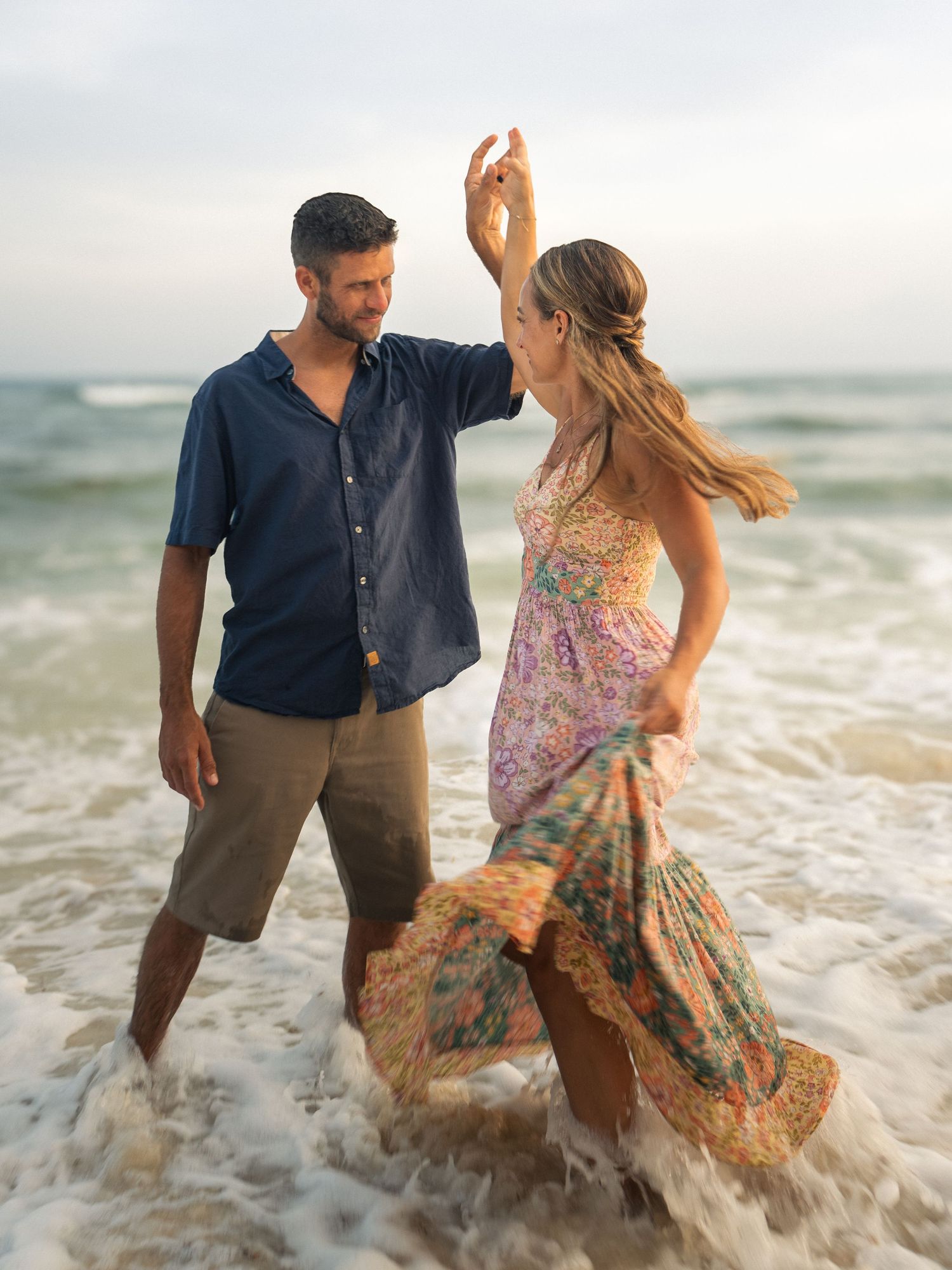 Romantic couple dancing in ocean waves at sunset, man in blue shirt twirling woman in floral maxi dress.