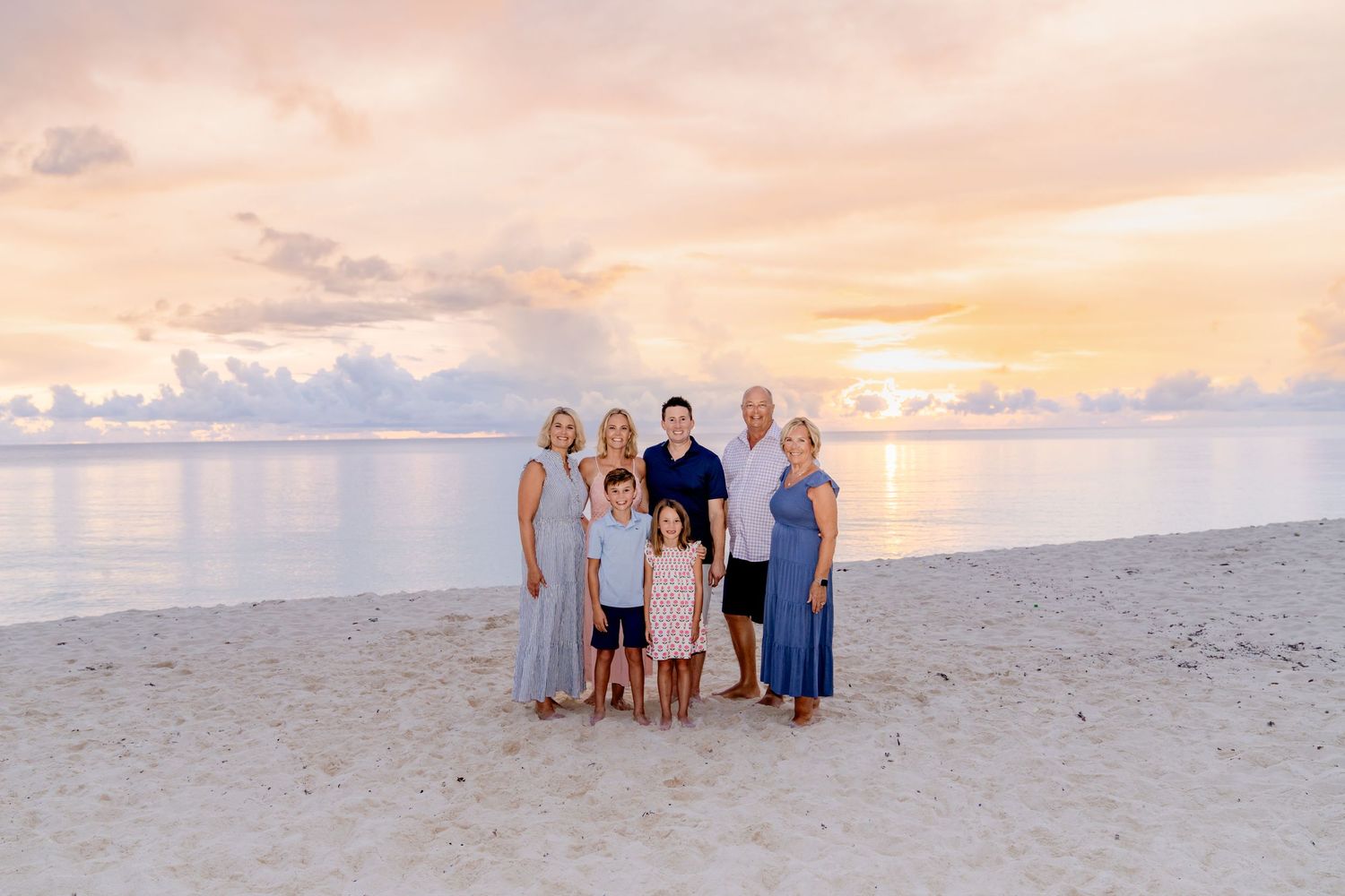 A group of people standing together on a sandy beach at sunset with pink and orange clouds in the sky.