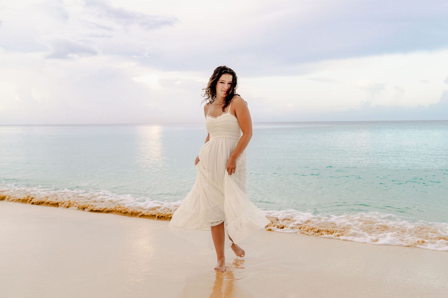A white dress flows elegantly against the backdrop of a serene beach landscape at sunset.