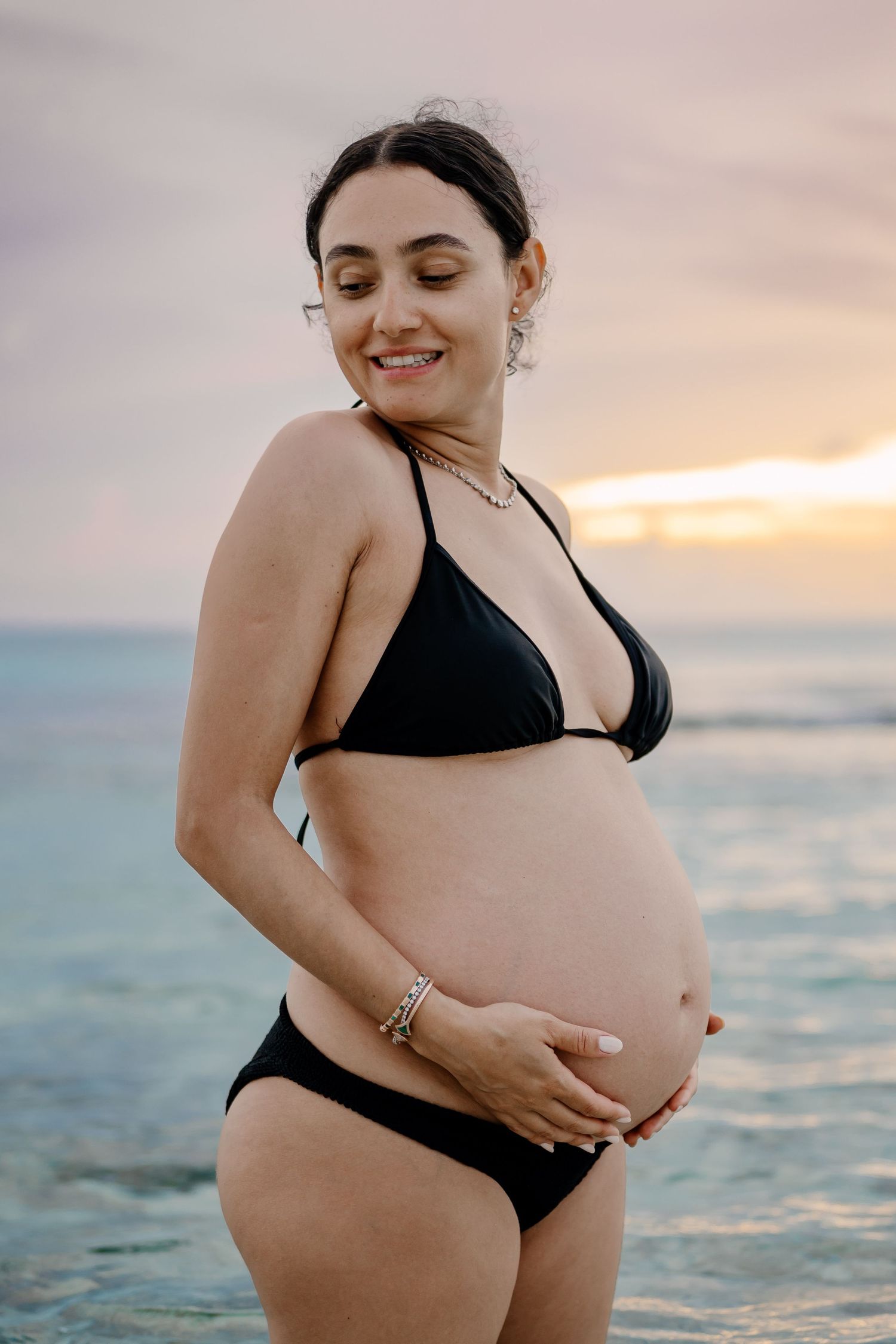 Sunset beach maternity session in black swimwear with gentle waves in background.