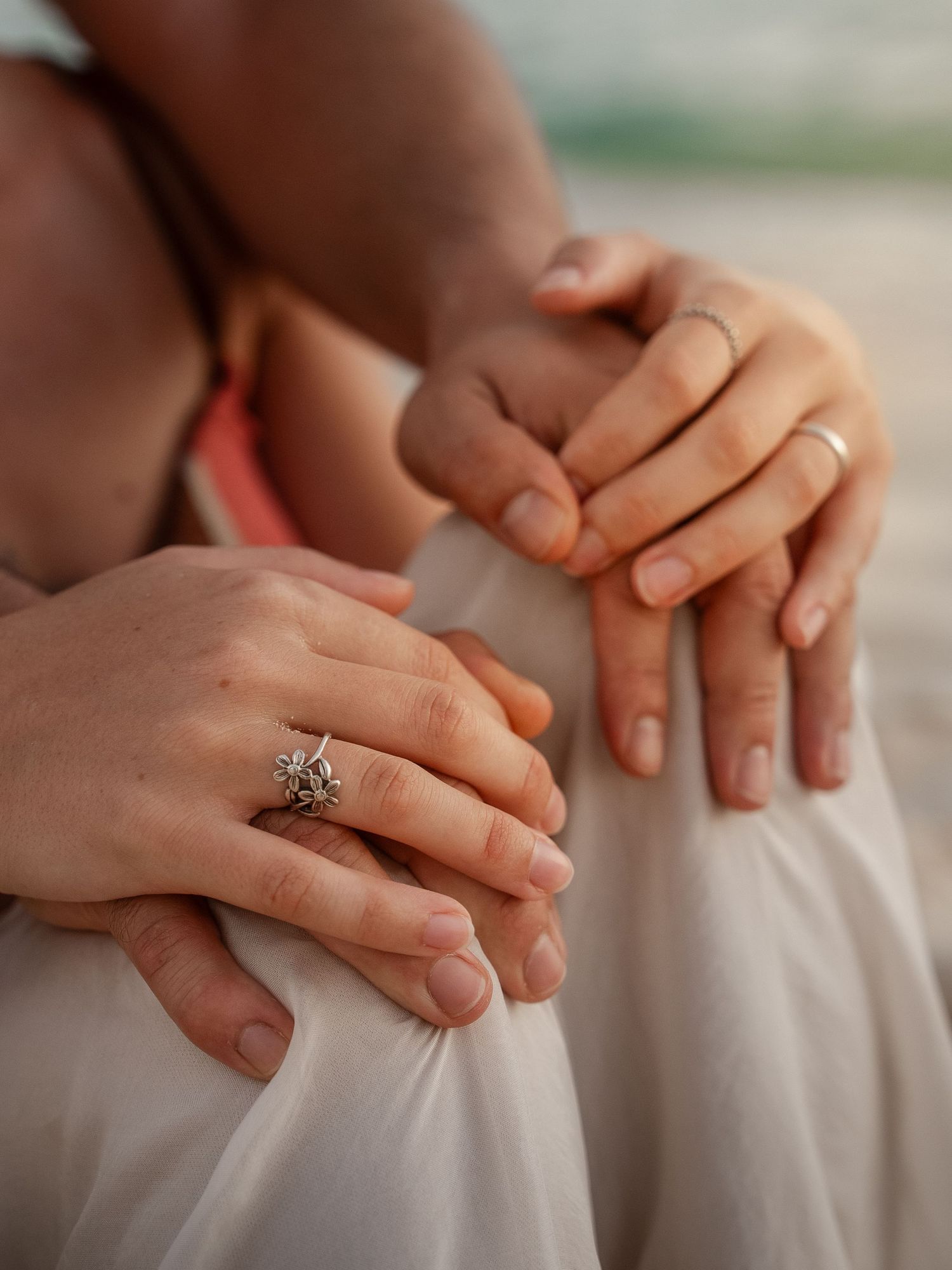 Close-up of couple's hands with elegant floral engagement ring and wedding band on beach.