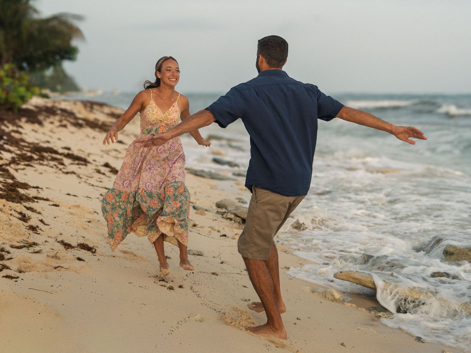 Couple dancing playfully on a tropical beach at sunset, woman in floral dress and man in navy shirt.