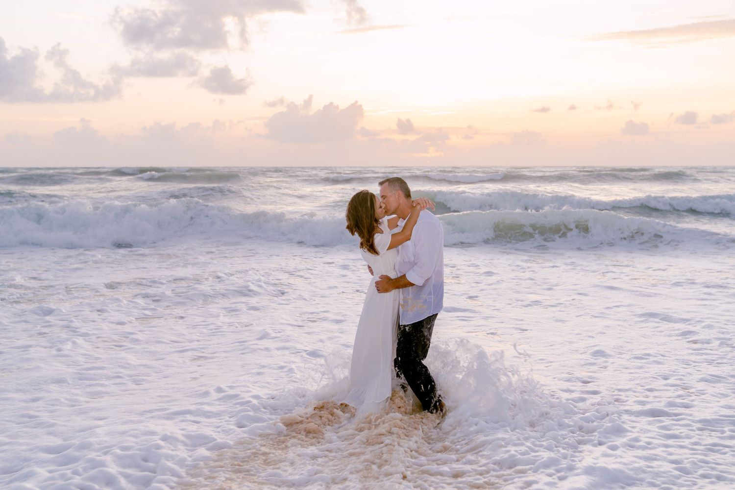 Couple embracing in ocean waves during golden hour beach photoshoot.