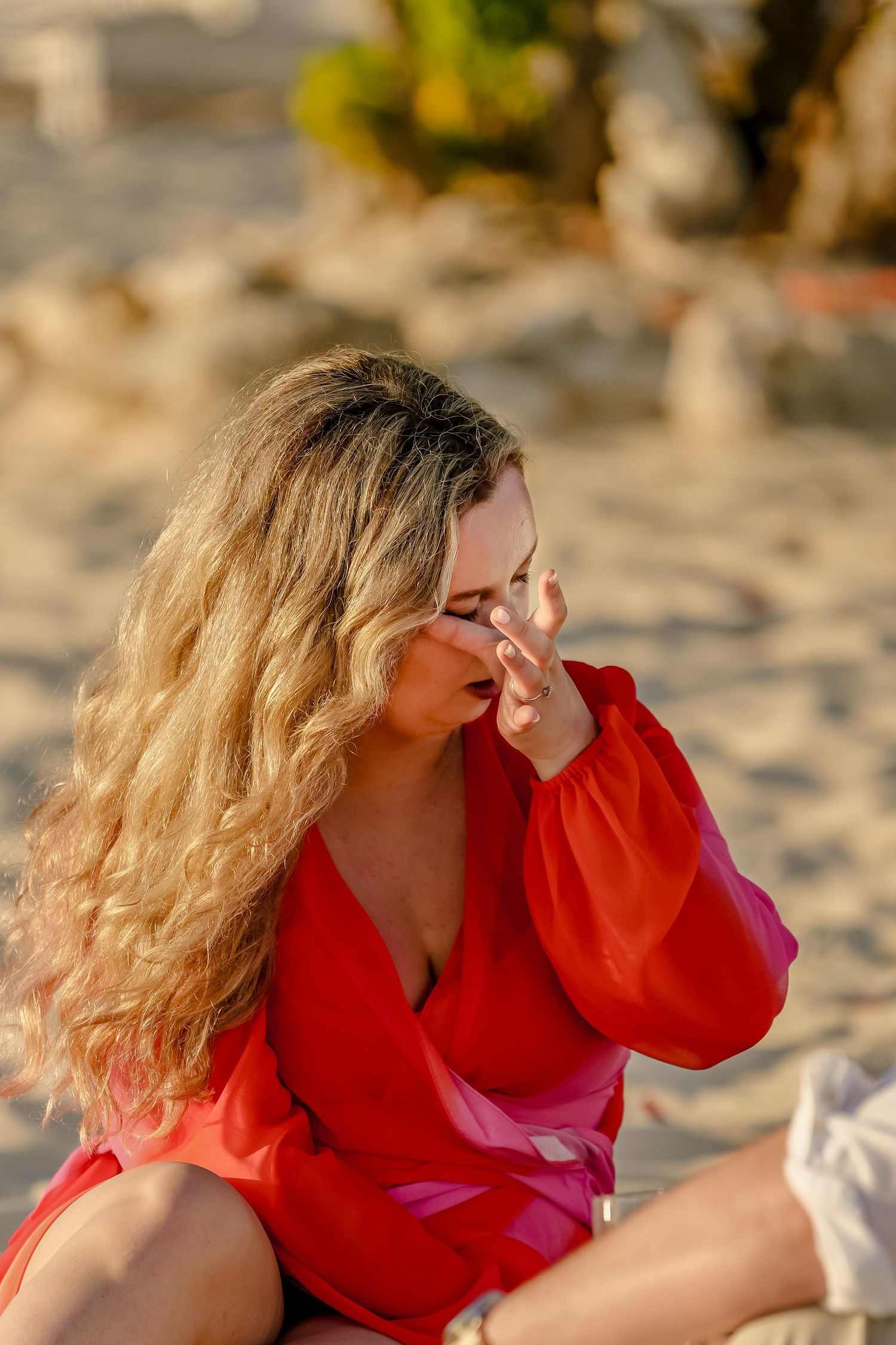 A woman in a bright red dress sits on a sandy beach during golden hour.