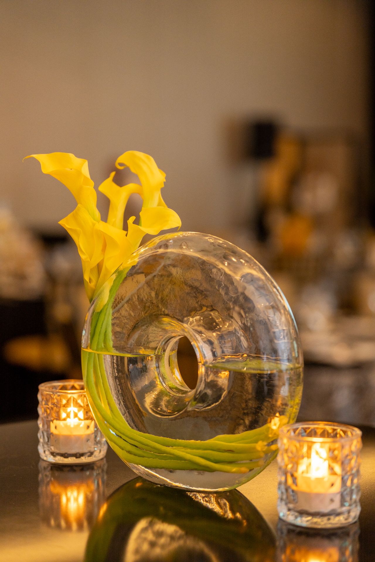 Yellow calla lilies in round glass vase with glowing tea light candles for elegant table decor.