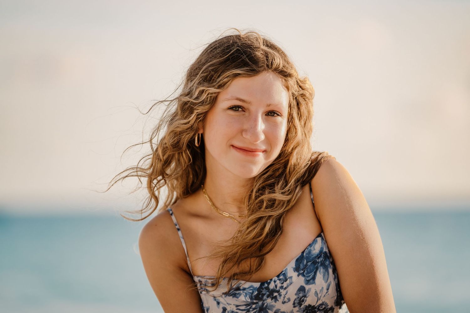 Person in floral dress smiling against beach backdrop during golden hour.